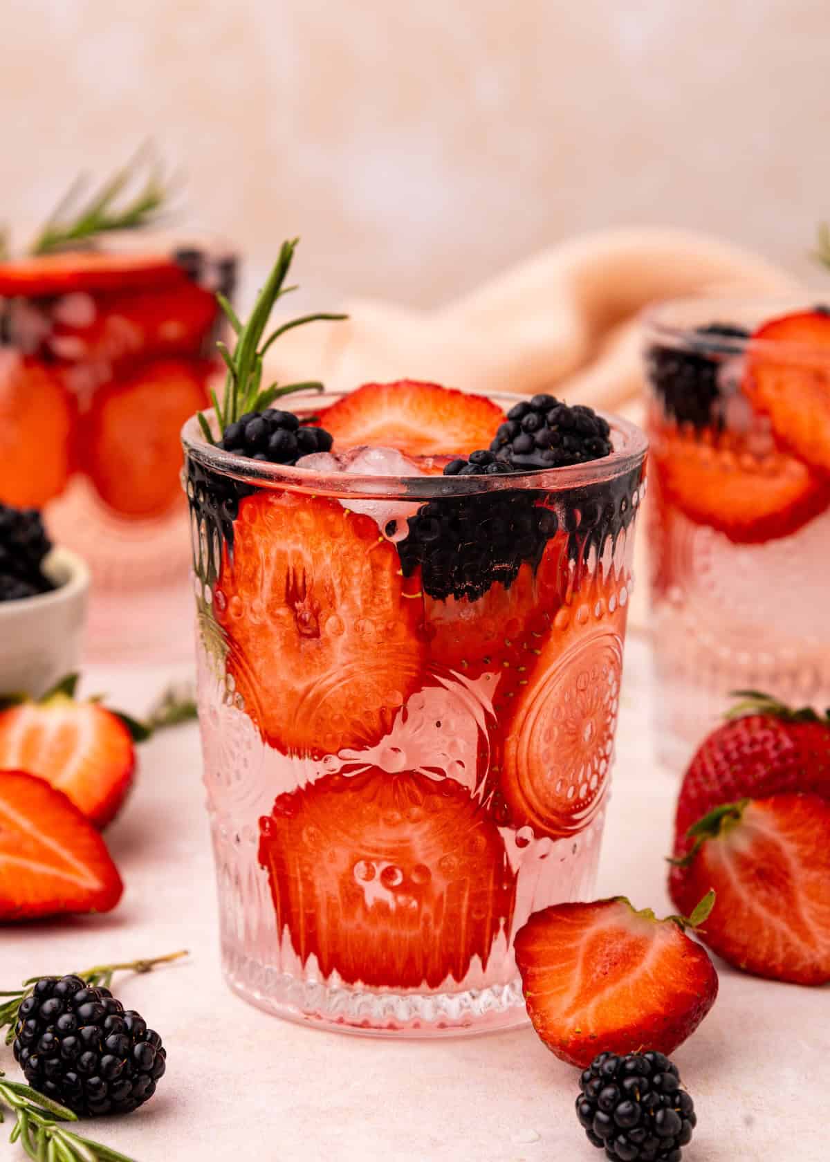A glass of strawberry water with ice, blackberries, and a fresh rosemary garnish.