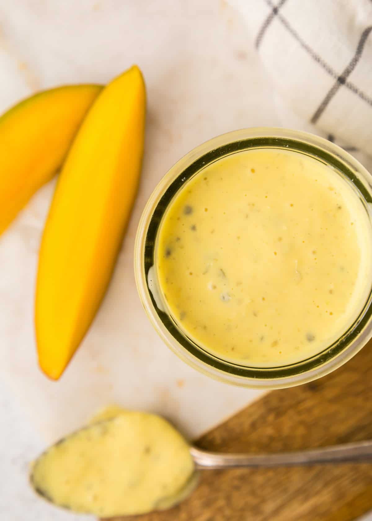 A jar of remoulade with a spoon and mango slices in the background.