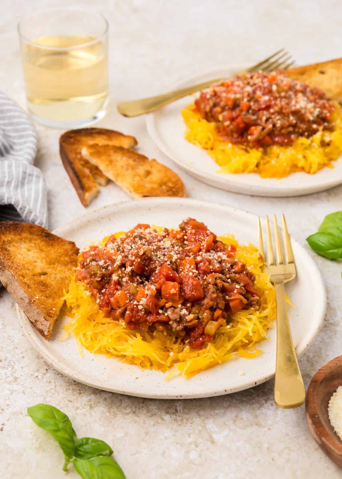 Two plates of spaghetti squash bolognese, toast, basil, and a glass of white wine.