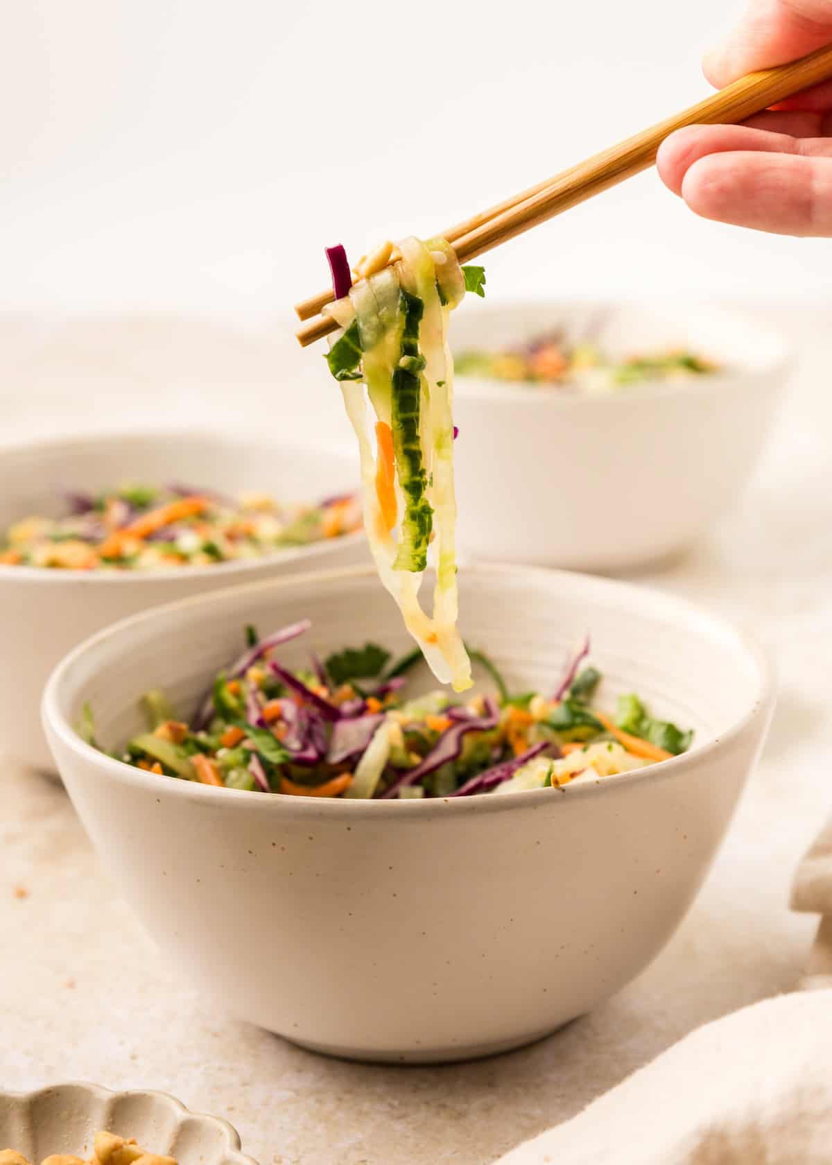 Chopsticks holding Asian Cucumber Noodle Salad over a white bowl of colorful veggies.