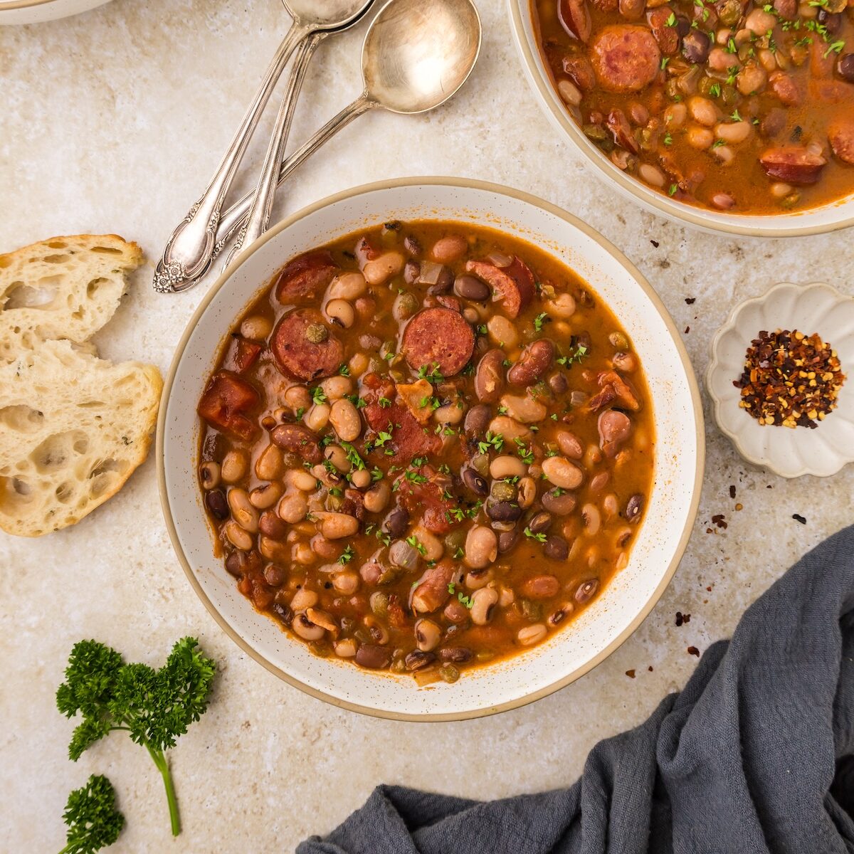 Bowls of hearty bean soup with sausage, bread, parsley, spoons, and red pepper flakes on the side.