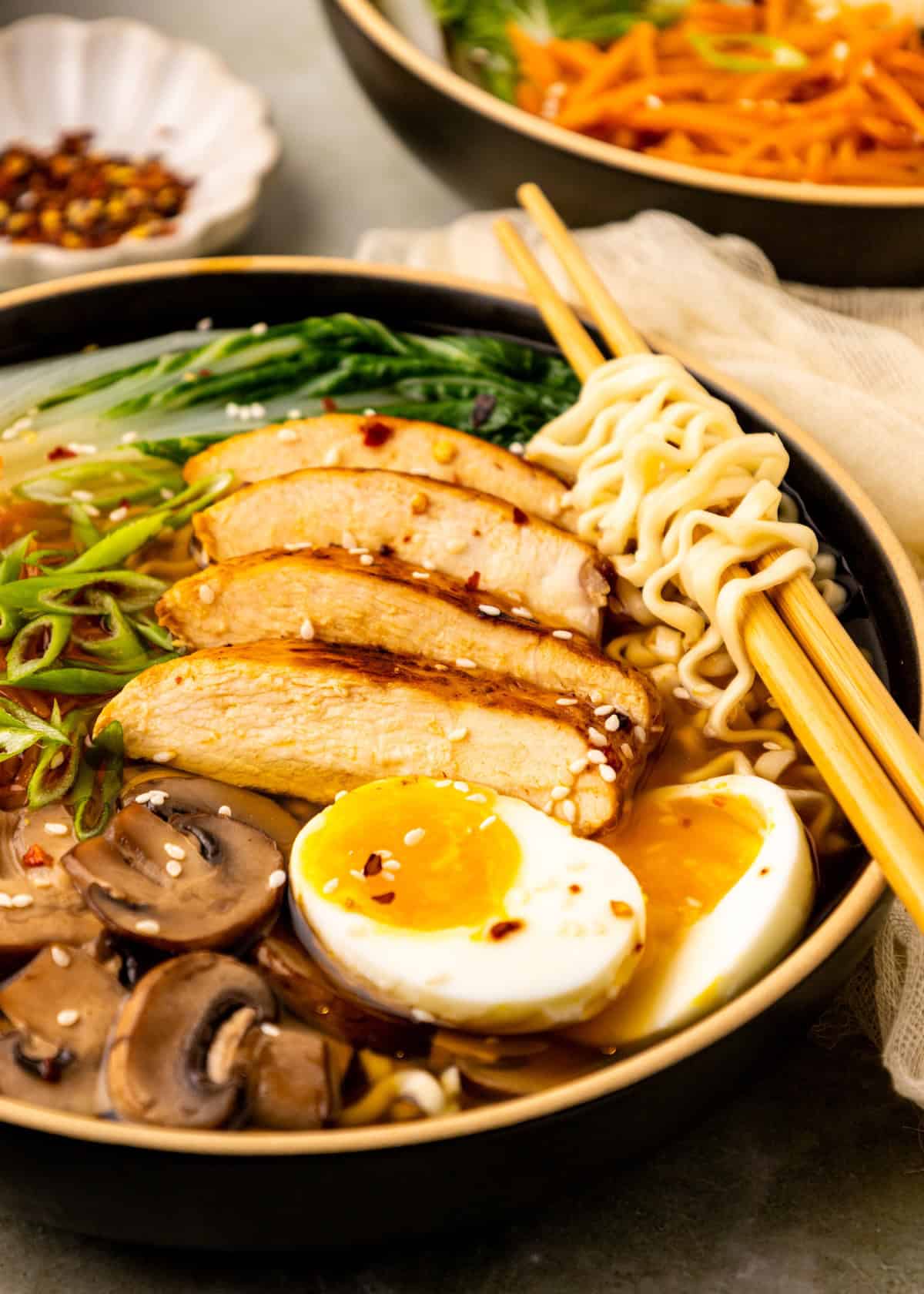 A bowl of Spicy Chicken Ramen with egg, mushrooms, greens, and chopsticks on top.