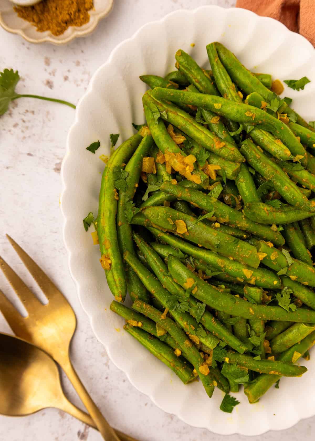 Curried green beans with chopped herbs in a white bowl, served beside gold utensils.