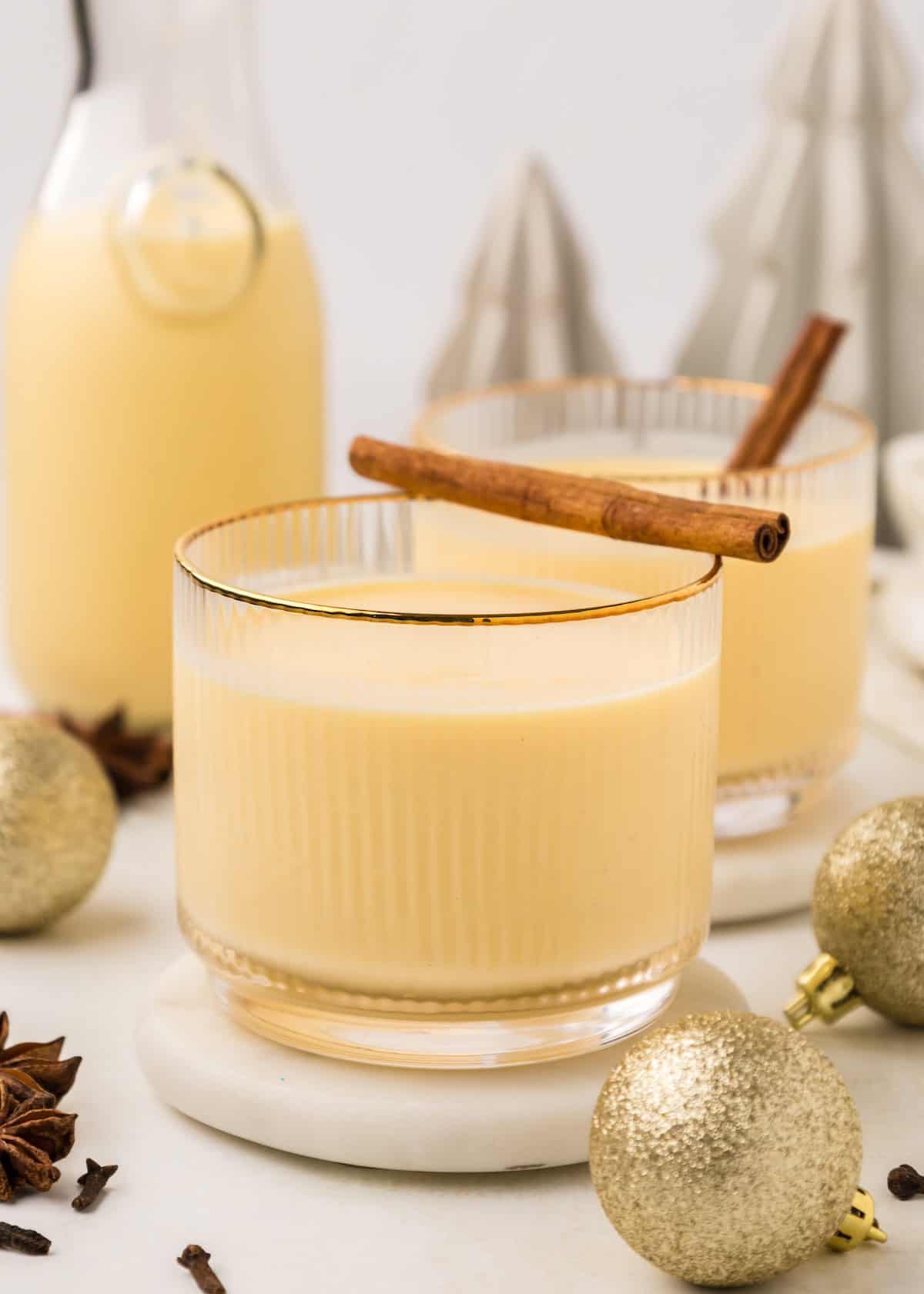 Glass of homemade eggnog with a cinnamon stick, surrounded by gold ornaments and festive decor.