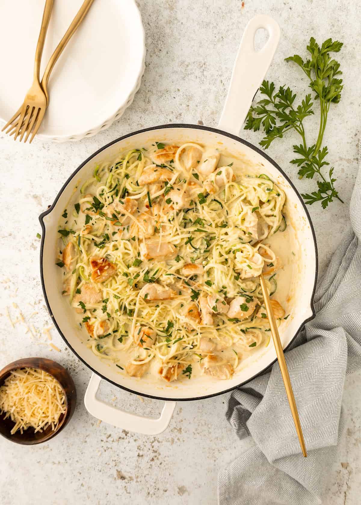 Creamy Chicken Zucchini Noodles in a white skillet, topped with herbs and cheese, plates nearby.