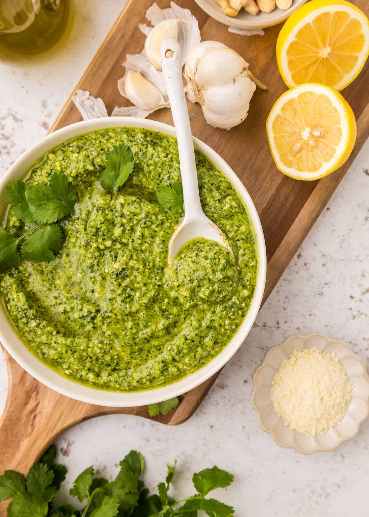 Bowl of cilantro pesto with a spoon, surrounded by lemons, garlic, cheese, and herbs.
