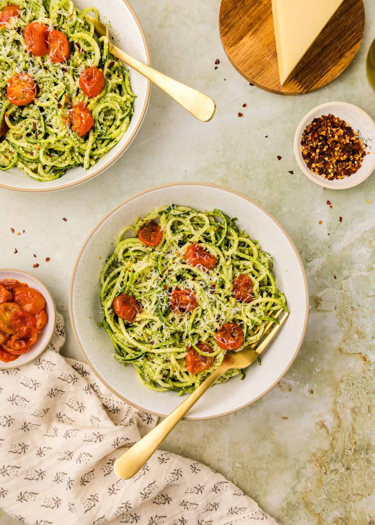 Two bowls of pesto zucchini noodles with cherry tomatoes and cheese, served with gold forks.