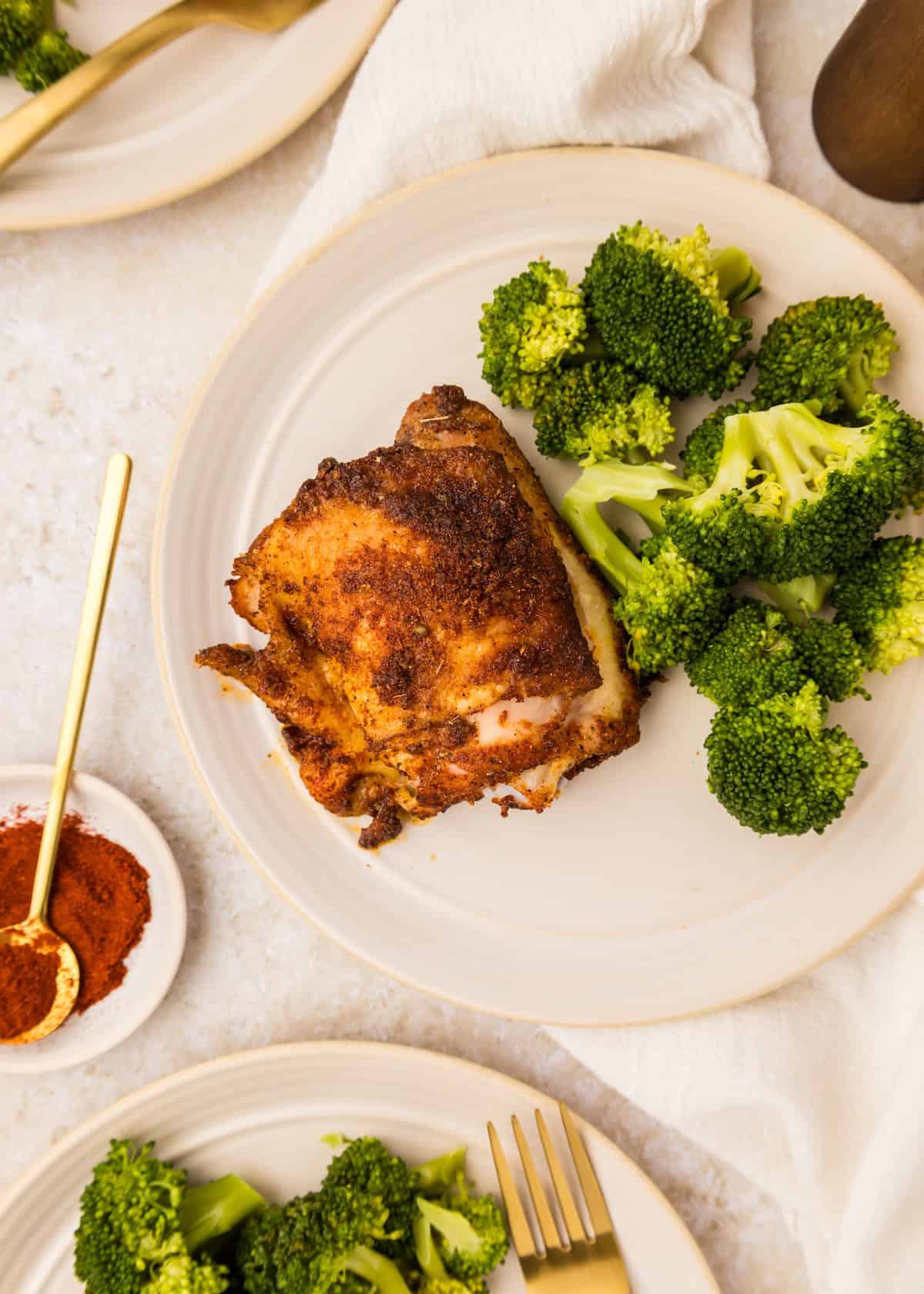 A plate with paprika chicken thigh and steamed broccoli on a light-colored surface.
