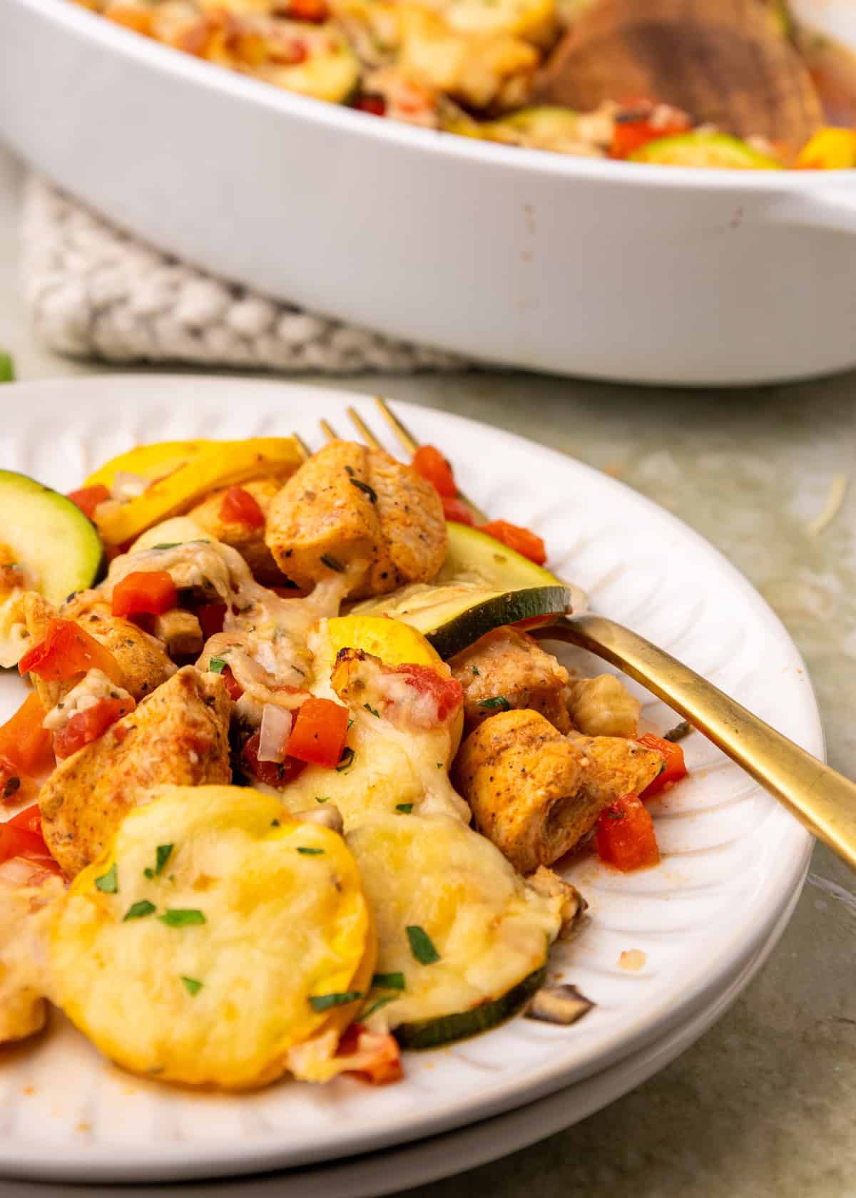 A plate of cheesy Ratatouille Chicken Casserole with vegetables, fork, and casserole dish behind.