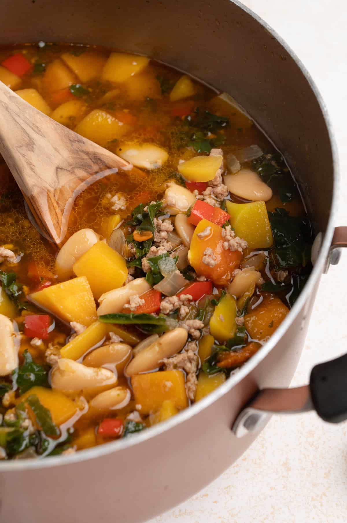 A pot of colorful pork chili and vegetable soup with a wooden spoon.