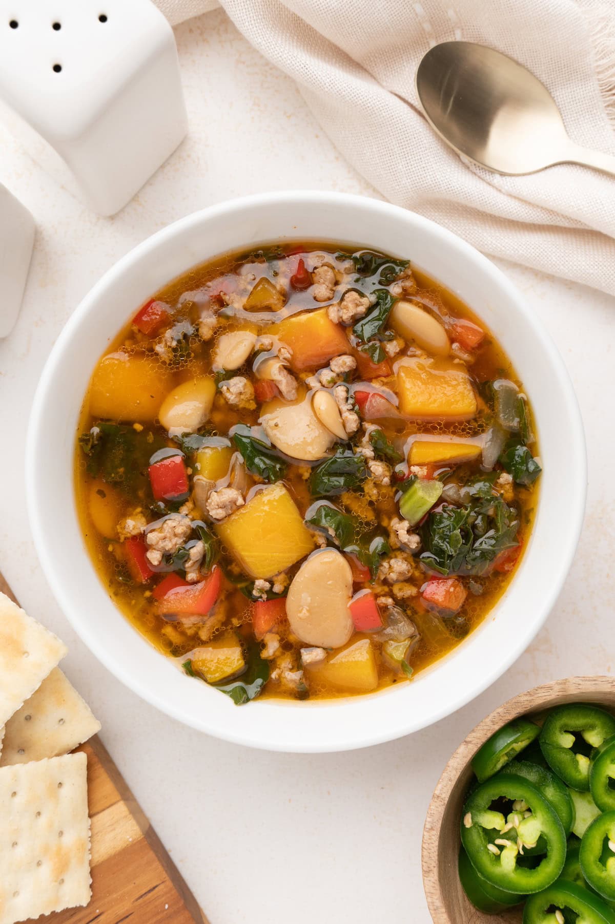 A bowl of pork chili soup with crackers and sliced jalapeños on the side.