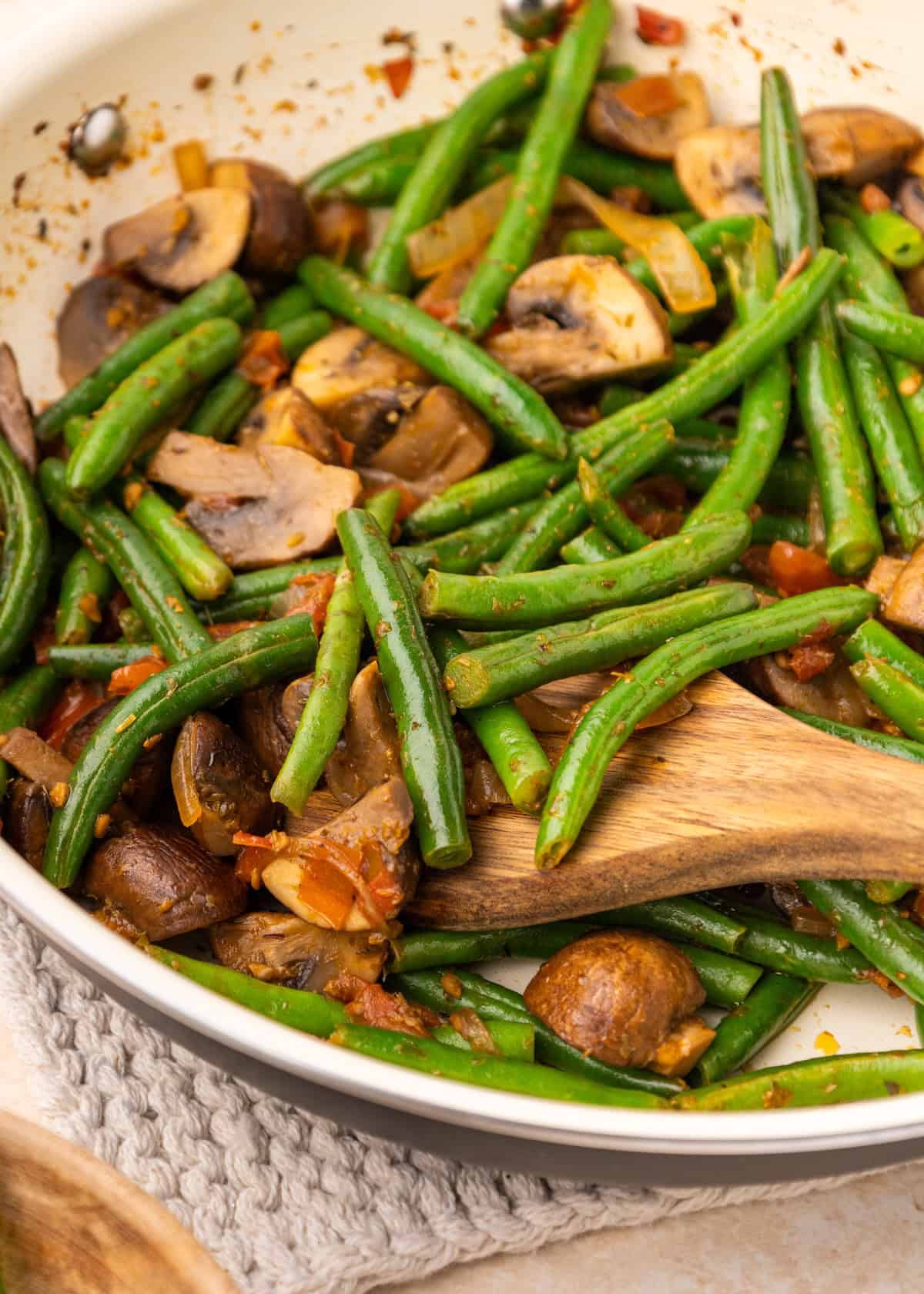 Sautéed green beans and mushrooms with tomatoes in a white skillet, stirred with a wooden spoon.