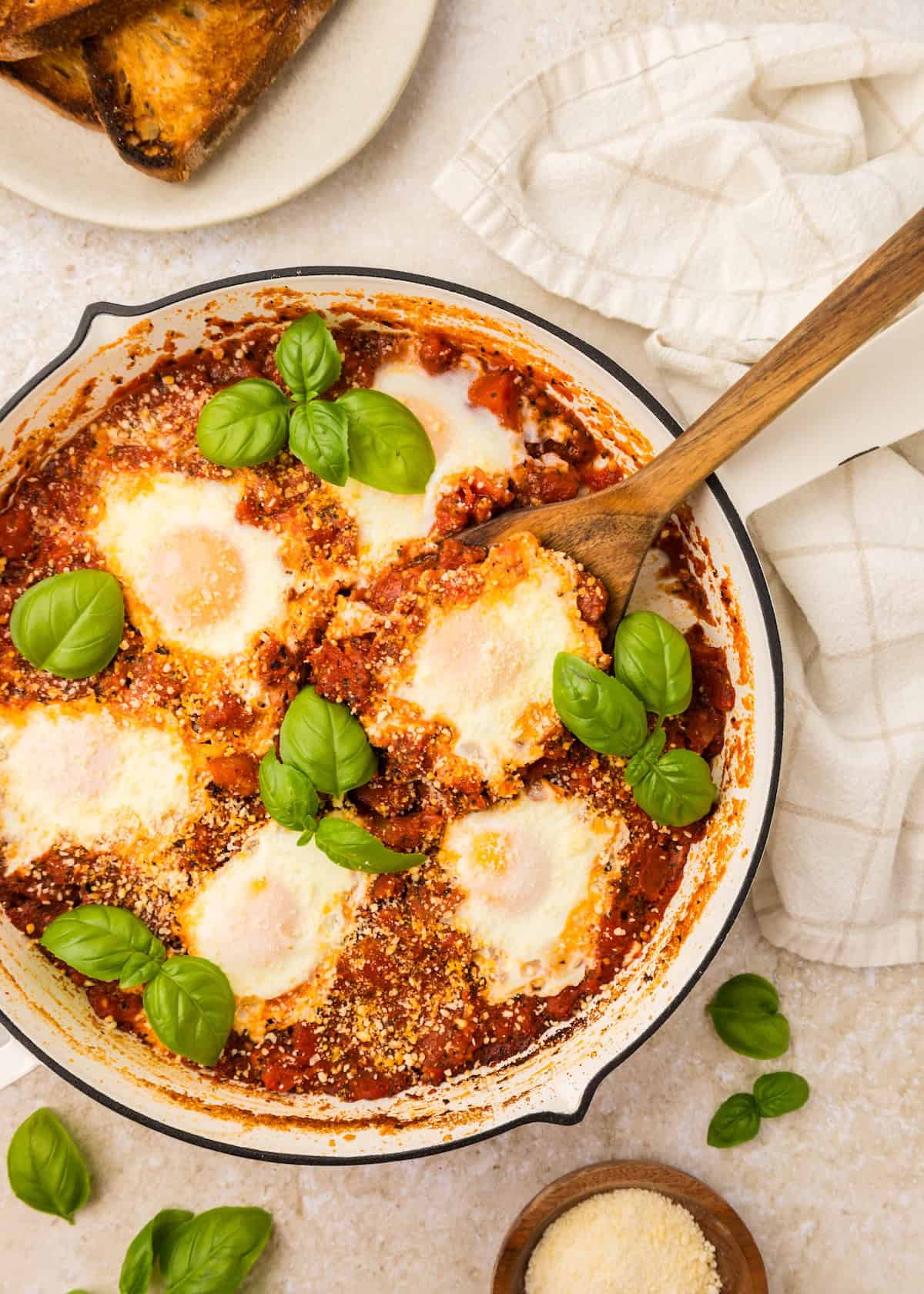Skillet with eggs in purgatory, topped with basil, served beside toast and a white towel.