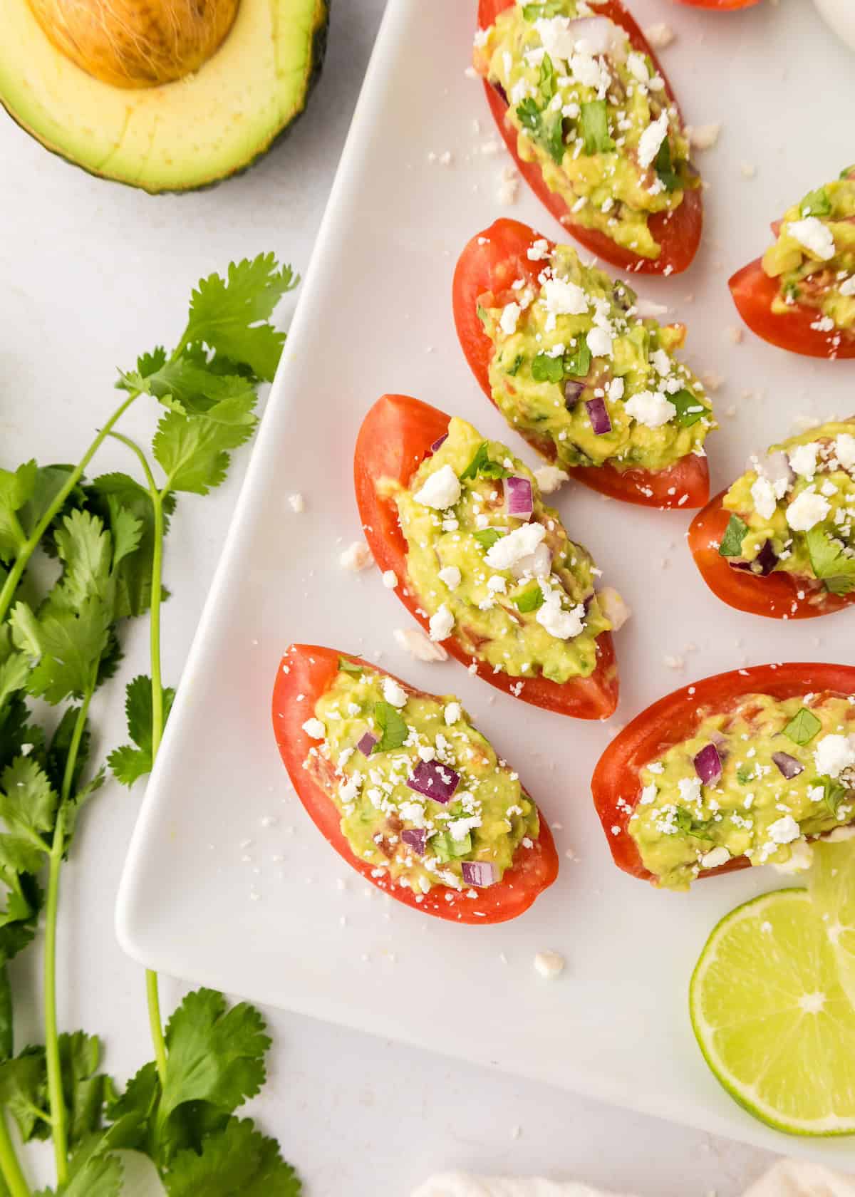 Guacamole bites: Tomato halves filled with guacamole, cheese, cilantro, and lime on a white plate.