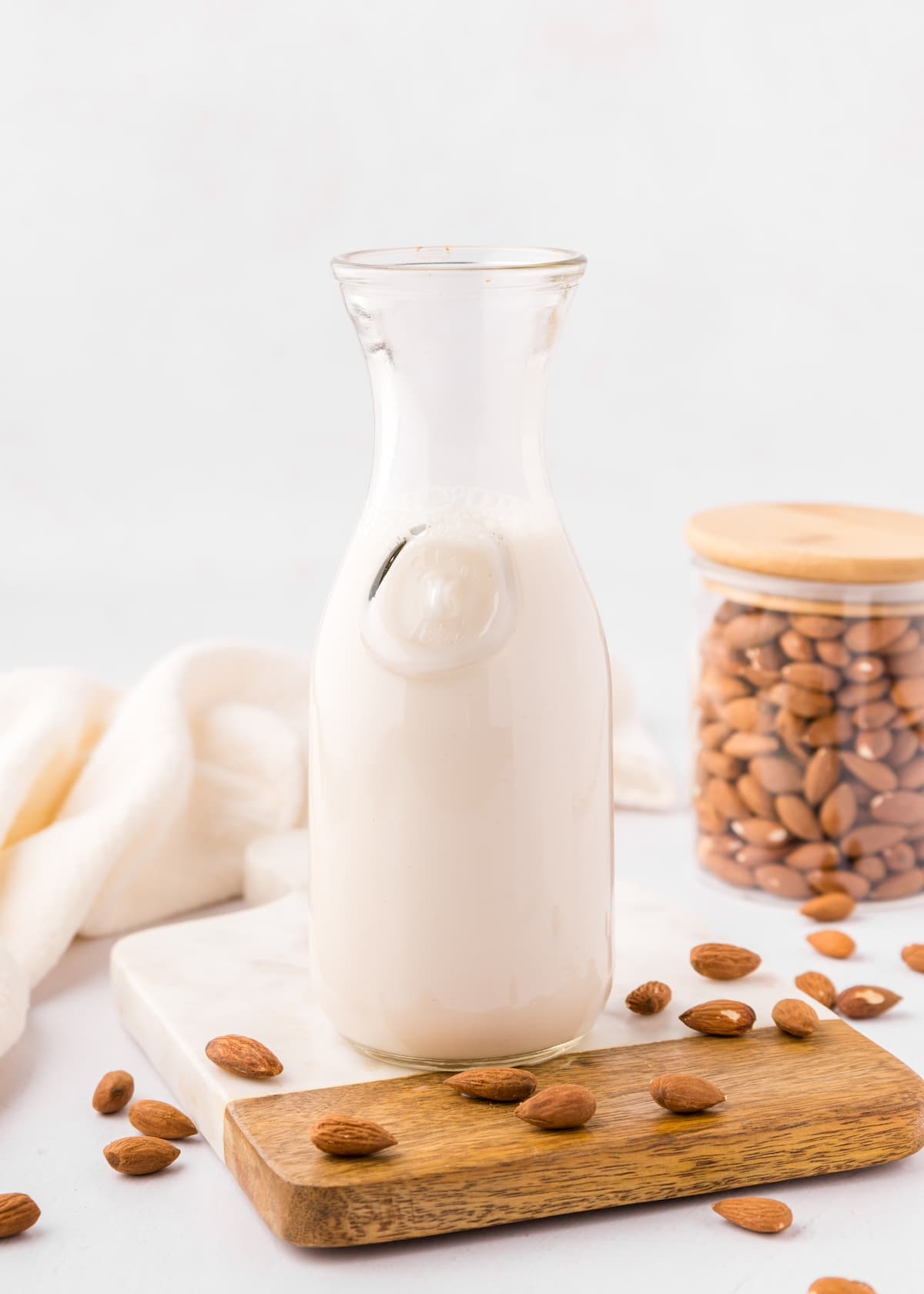 A glass carafe of homemade almond milk with almonds and a jar of almonds on a wooden board.