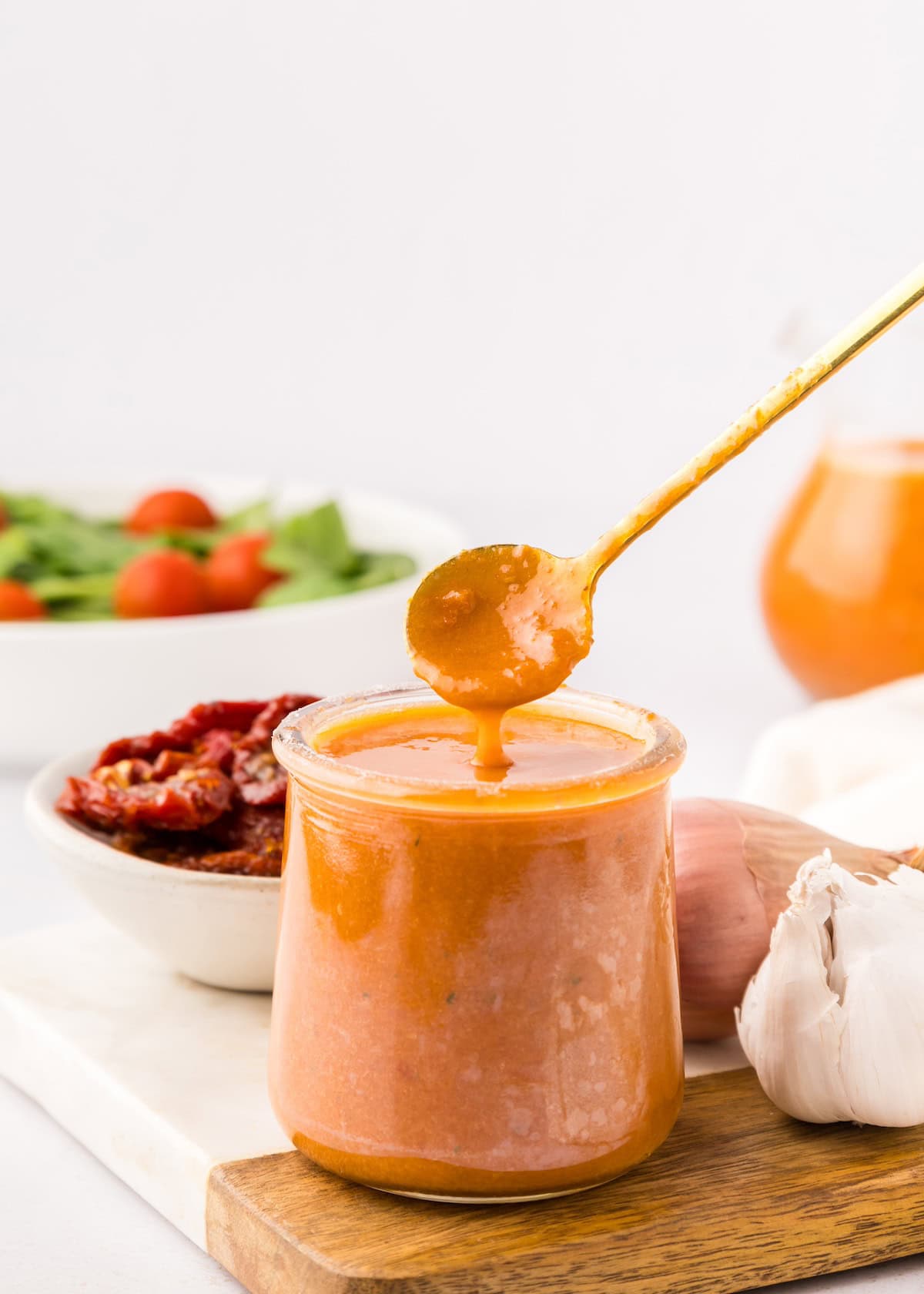 A jar of creamy sun-dried tomato vinaigrette with garlic, salad, and a spoon nearby.