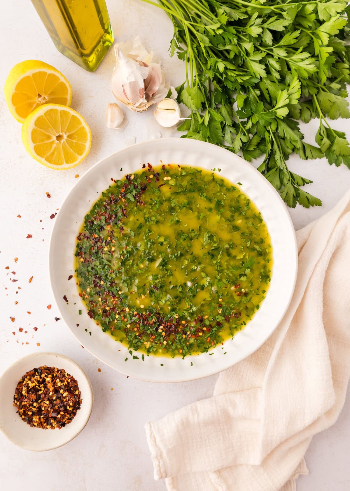 Chimichurri Sauce in a bowl, surrounded by parsley, lemon, garlic, and chili flakes.