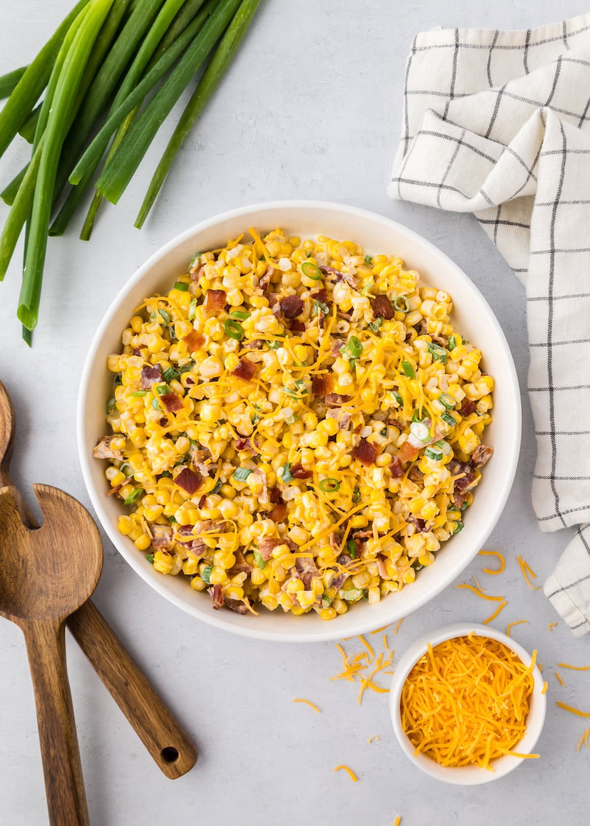A bowl of loaded sweet corn salad with bacon, cheese, green onions, and utensils nearby.