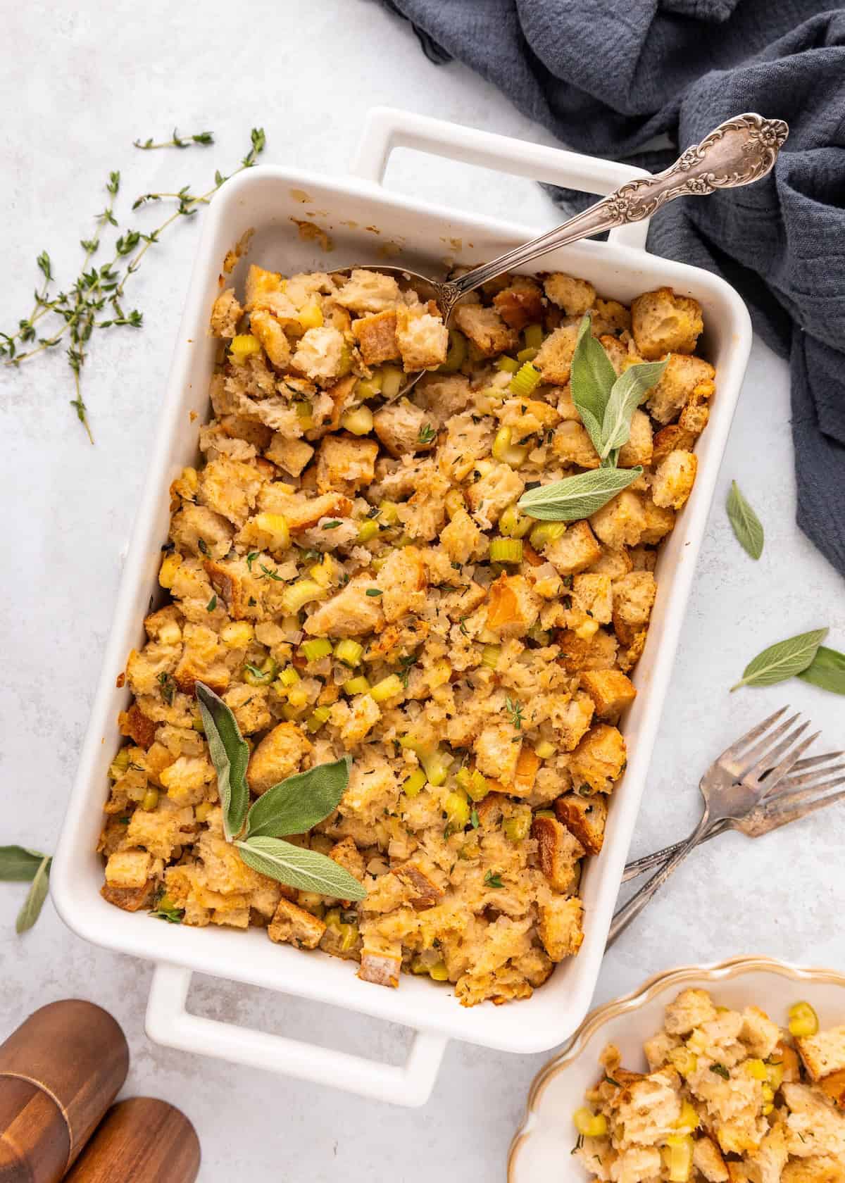 A casserole dish of sourdough stuffing, garnished with fresh sage and served with utensils.