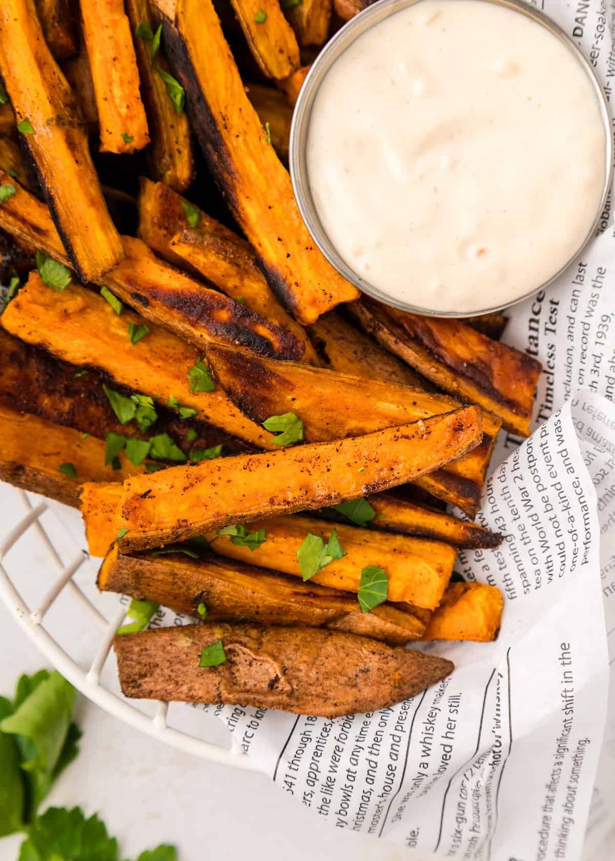 Sweet potato fries served on newspaper with dipping sauce and fresh herbs sprinkled on top.