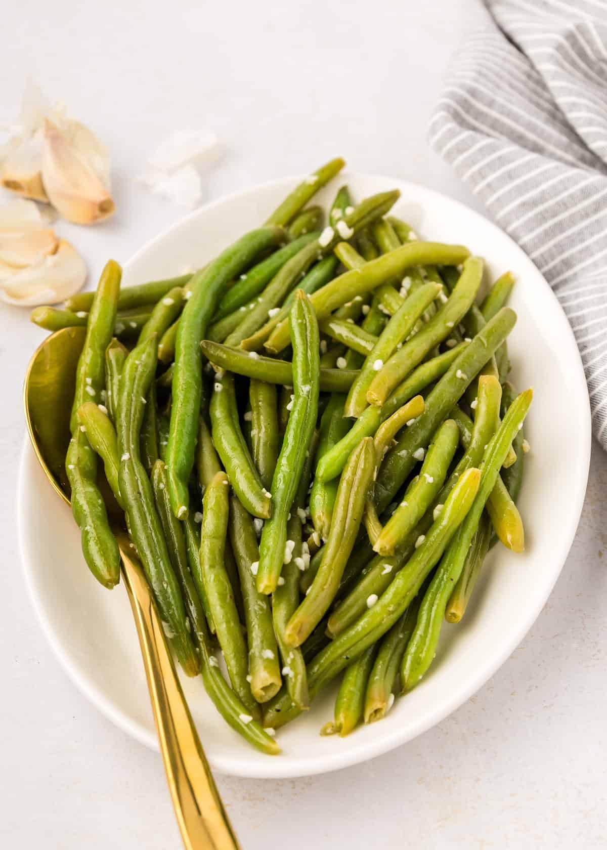 Oval white bowl of garlic green beans with sesame seeds, gold spoon, and a kitchen towel nearby.