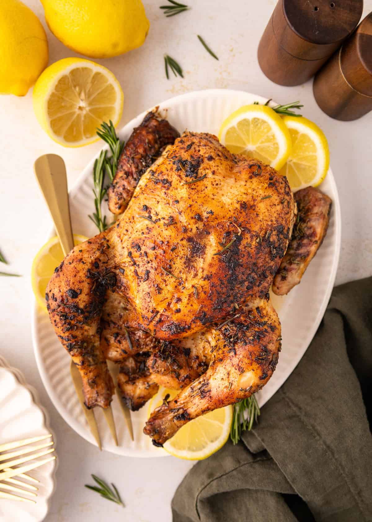 Lemon Rosemary Chicken on a platter with lemon slices, herbs, and utensils nearby.