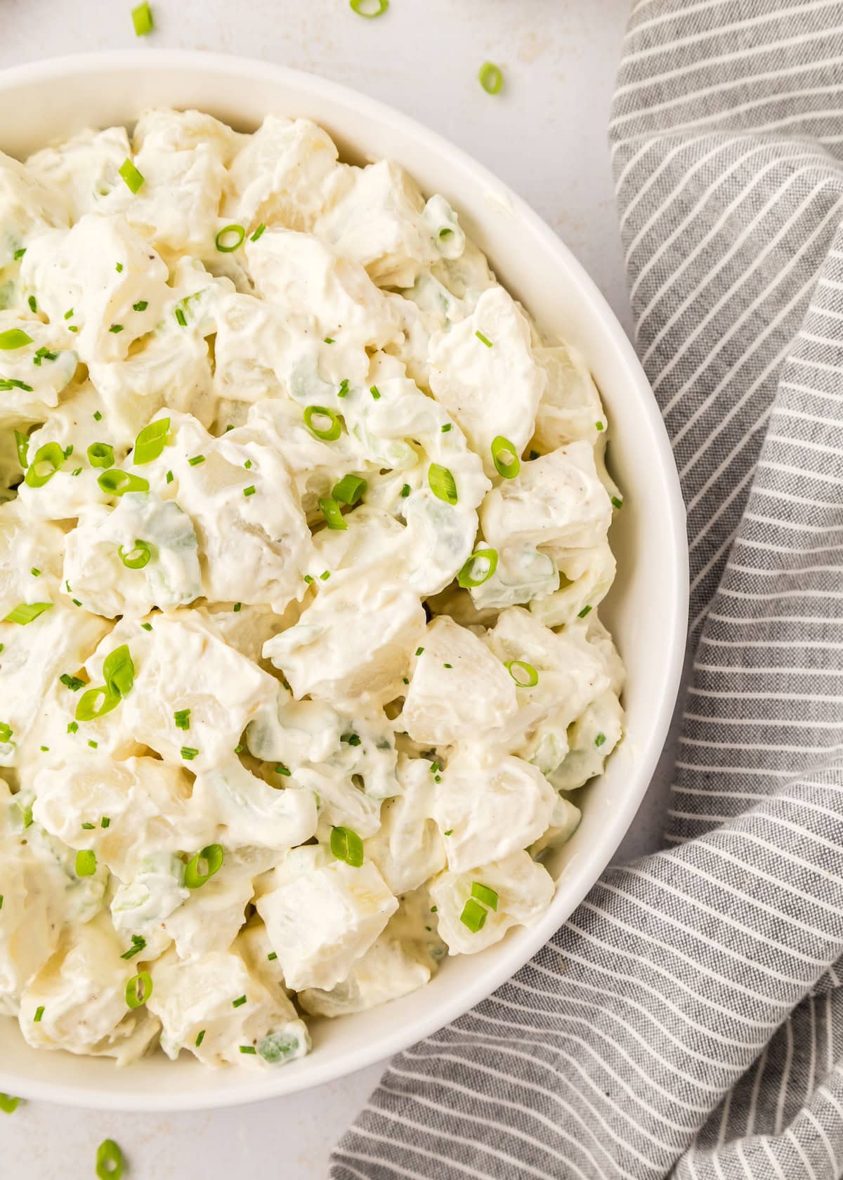 A bowl of Sour Cream Potato Salad topped with green onions sits by a striped gray napkin.