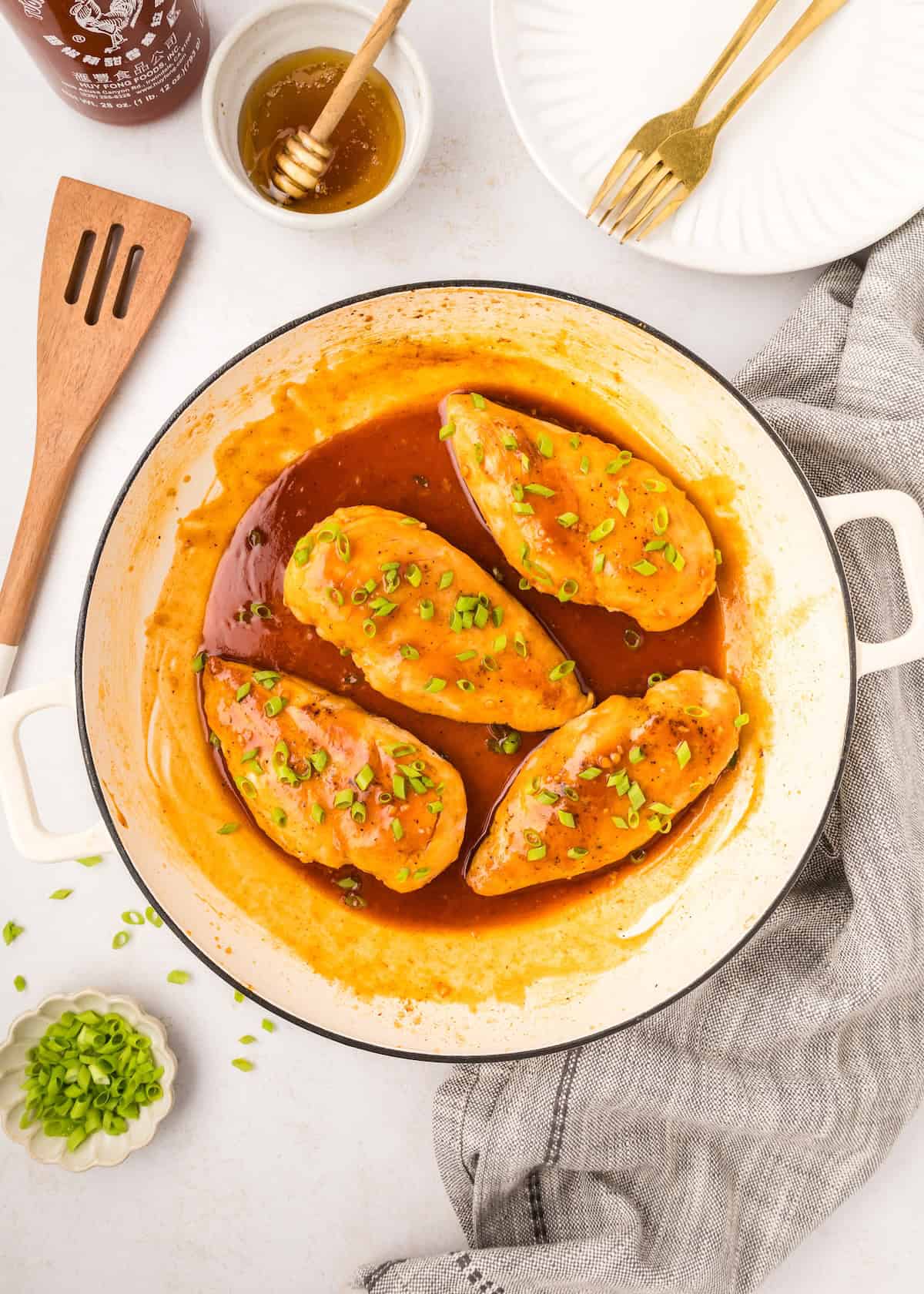 Four Honey Sriracha Chicken breasts with sauce and green onions in a white skillet.