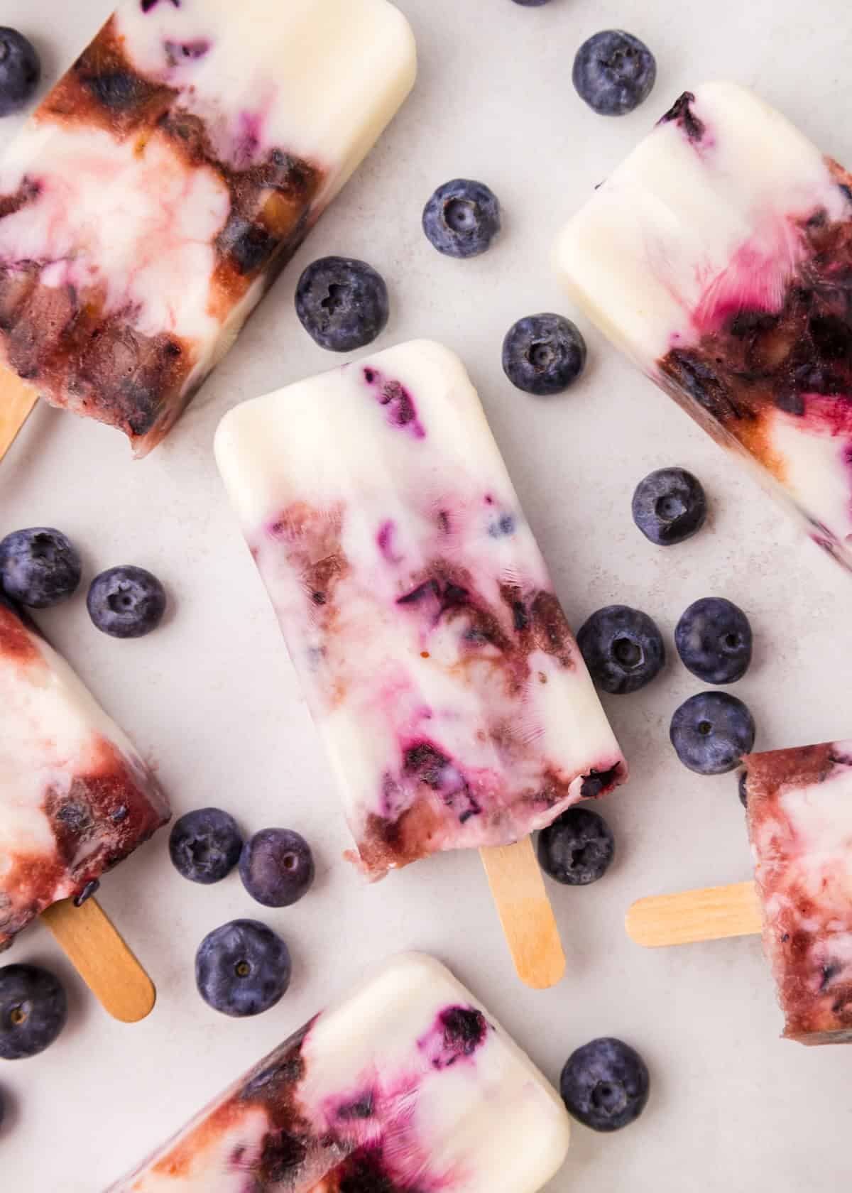 Blueberry yogurt popsicles surrounded by fresh blueberries on a white surface.