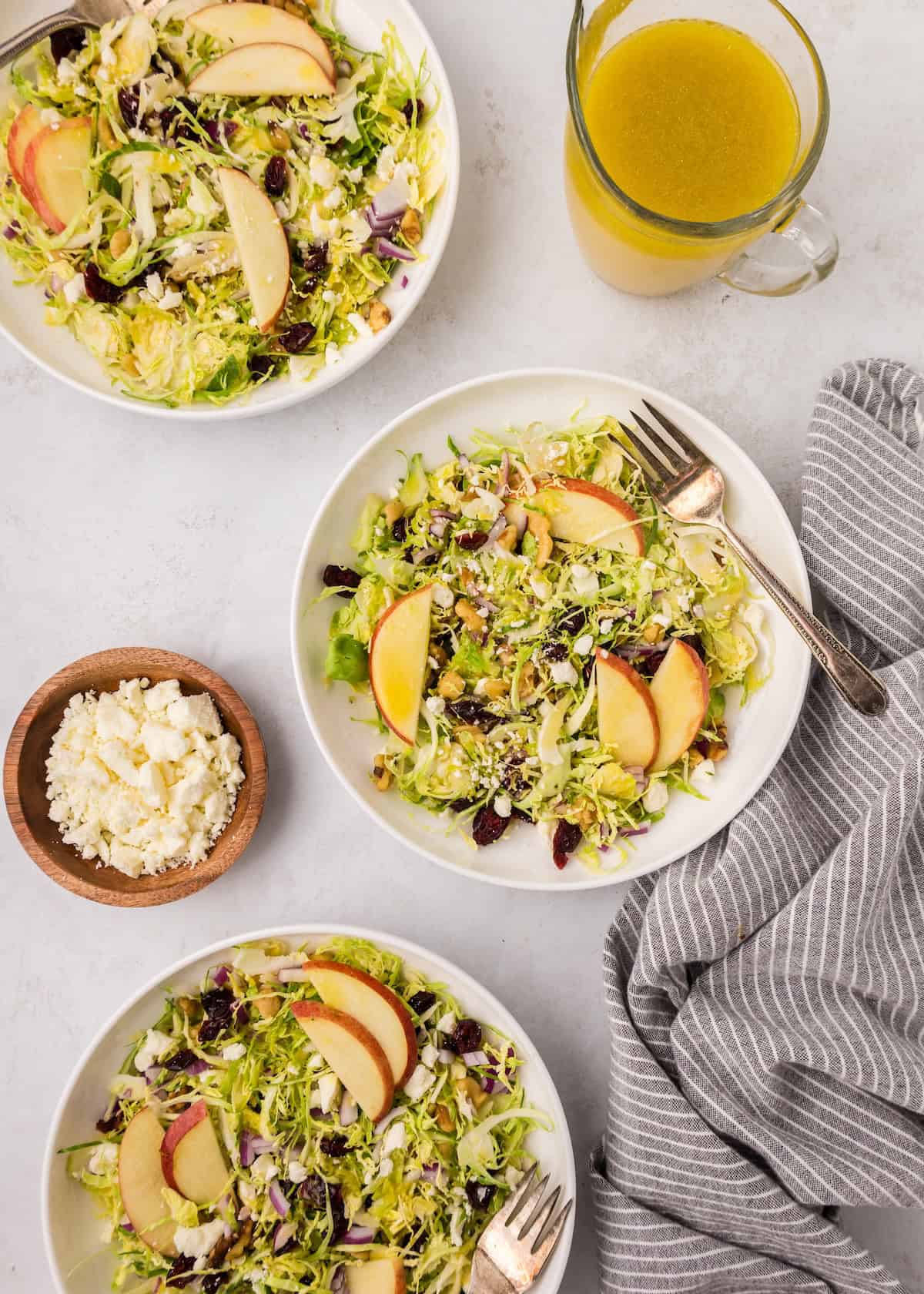 Three plates of Shaved Brussels Sprouts Salad with apple, feta, cranberries, and yellow dressing.