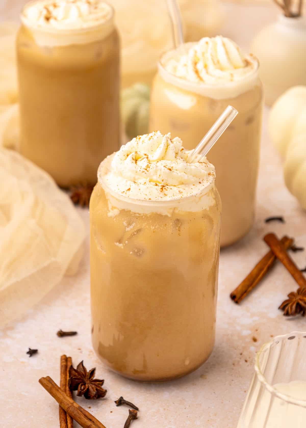 Iced Pumpkin Spice Lattes with whipped cream in glass jars, surrounded by spices and straws.