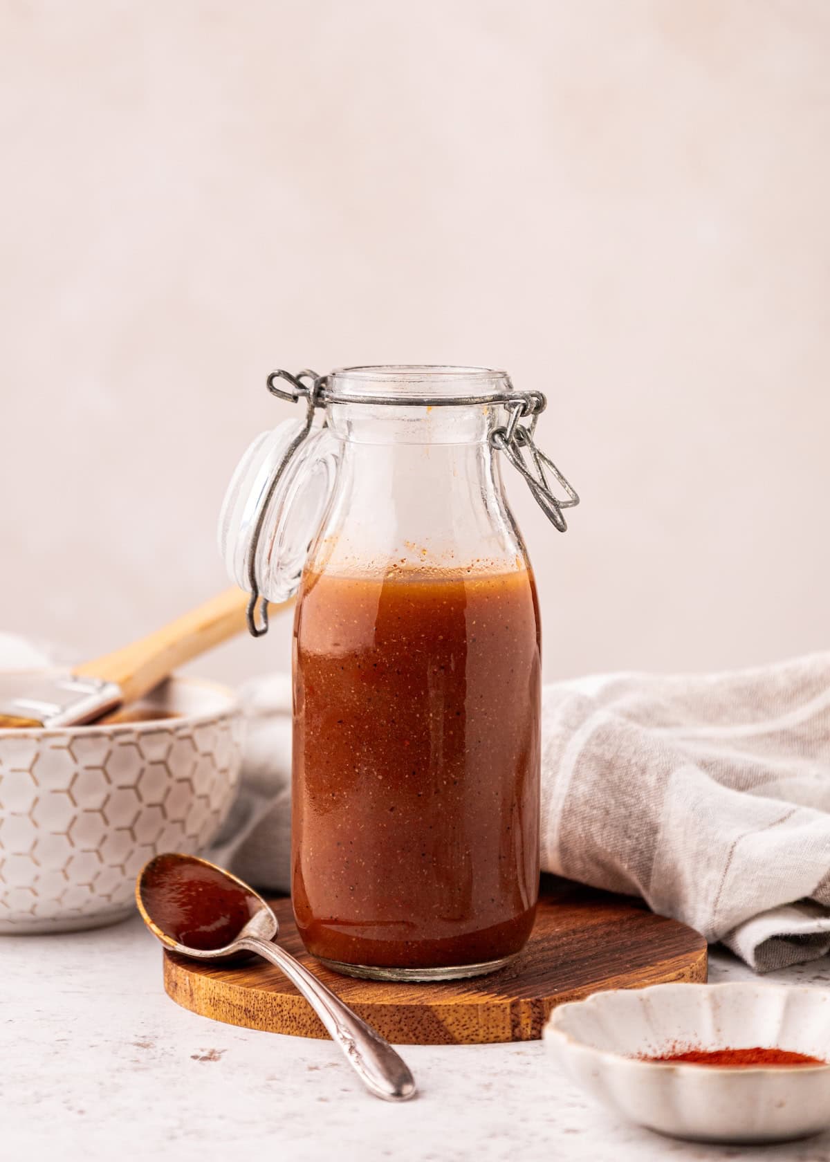 A glass jar of homemade BBQ sauce on a wooden coaster with a spoon and bowl nearby.