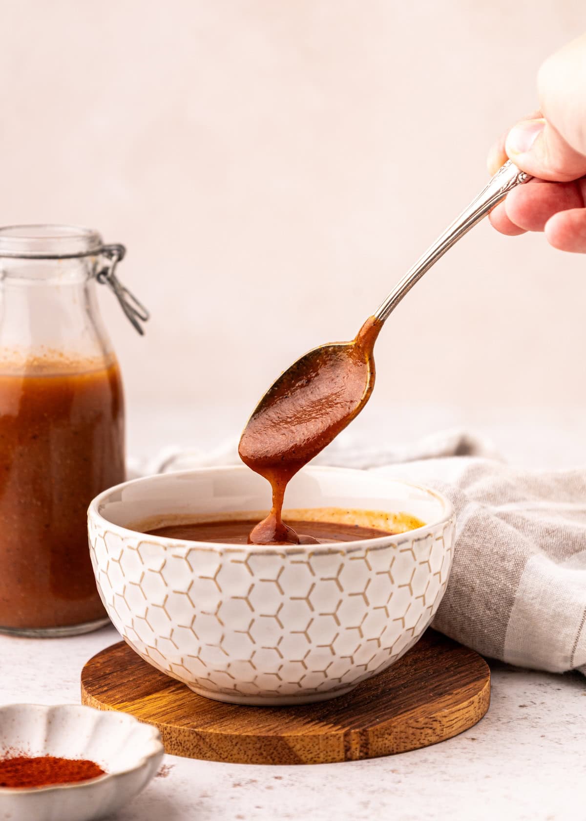 A hand drips homemade BBQ sauce from a spoon into a patterned bowl on a wooden coaster.