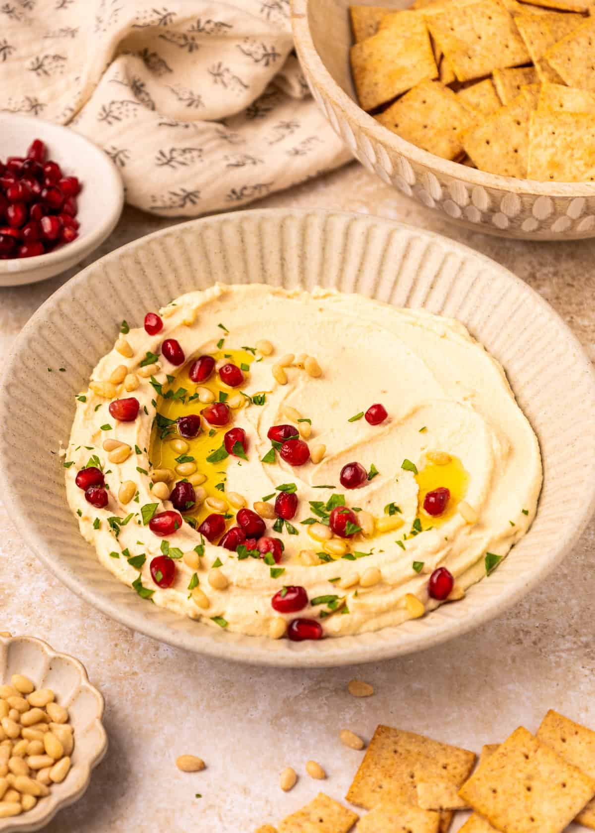 A bowl of Toasted Pine Nut Hummus topped with olive oil, pomegranate seeds, and herbs.