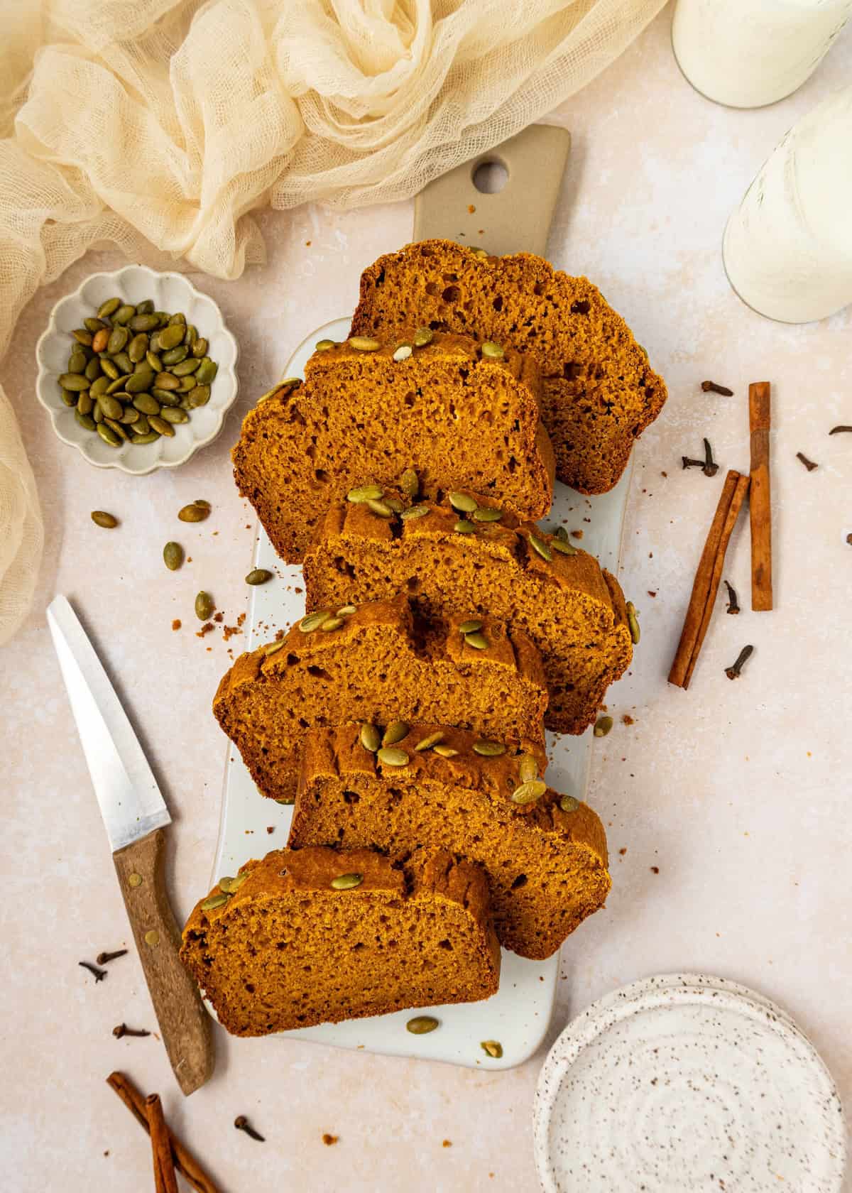 Pumpkin Bread slices with pumpkin seeds on a board, surrounded by spices and milk.