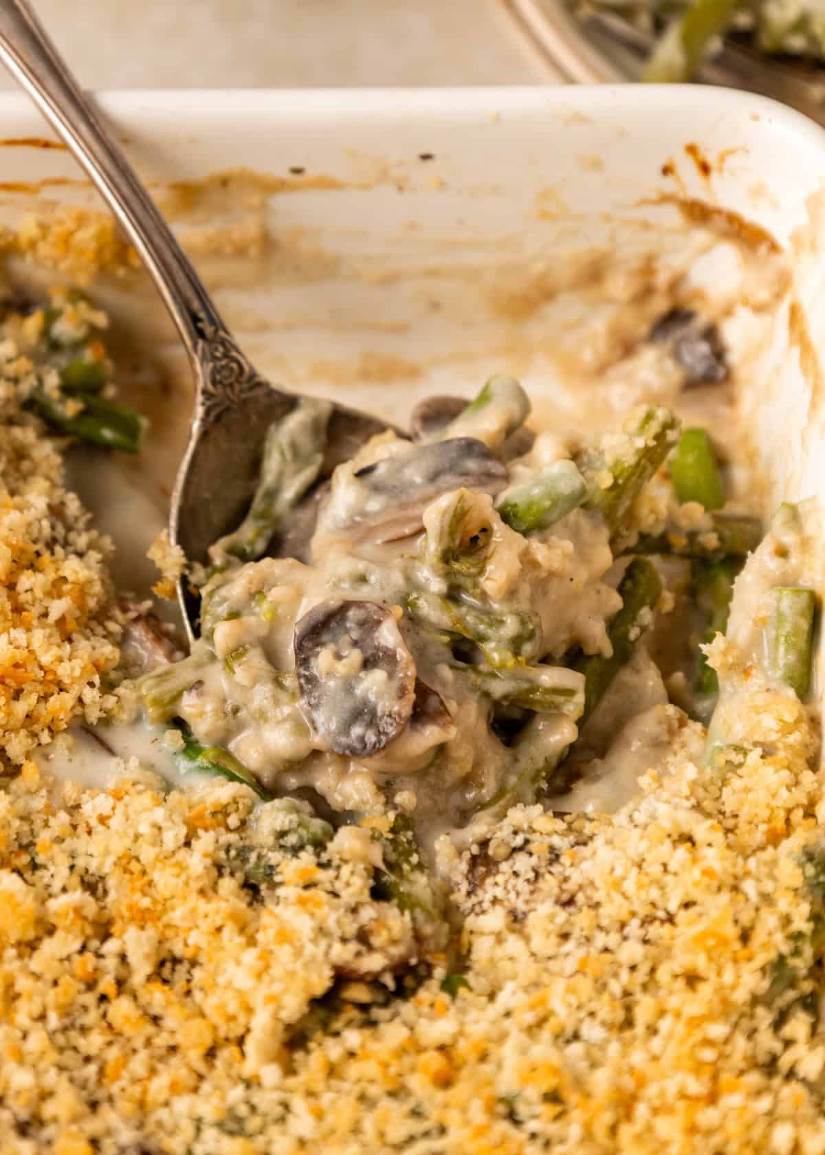 A spoon scoops creamy asparagus casserole with mushrooms and a crunchy breadcrumb topping.