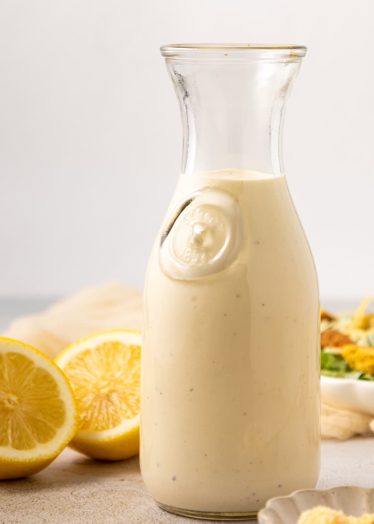 Glass bottle of Homemade Caesar Dressing with lemons in the background on a light surface.