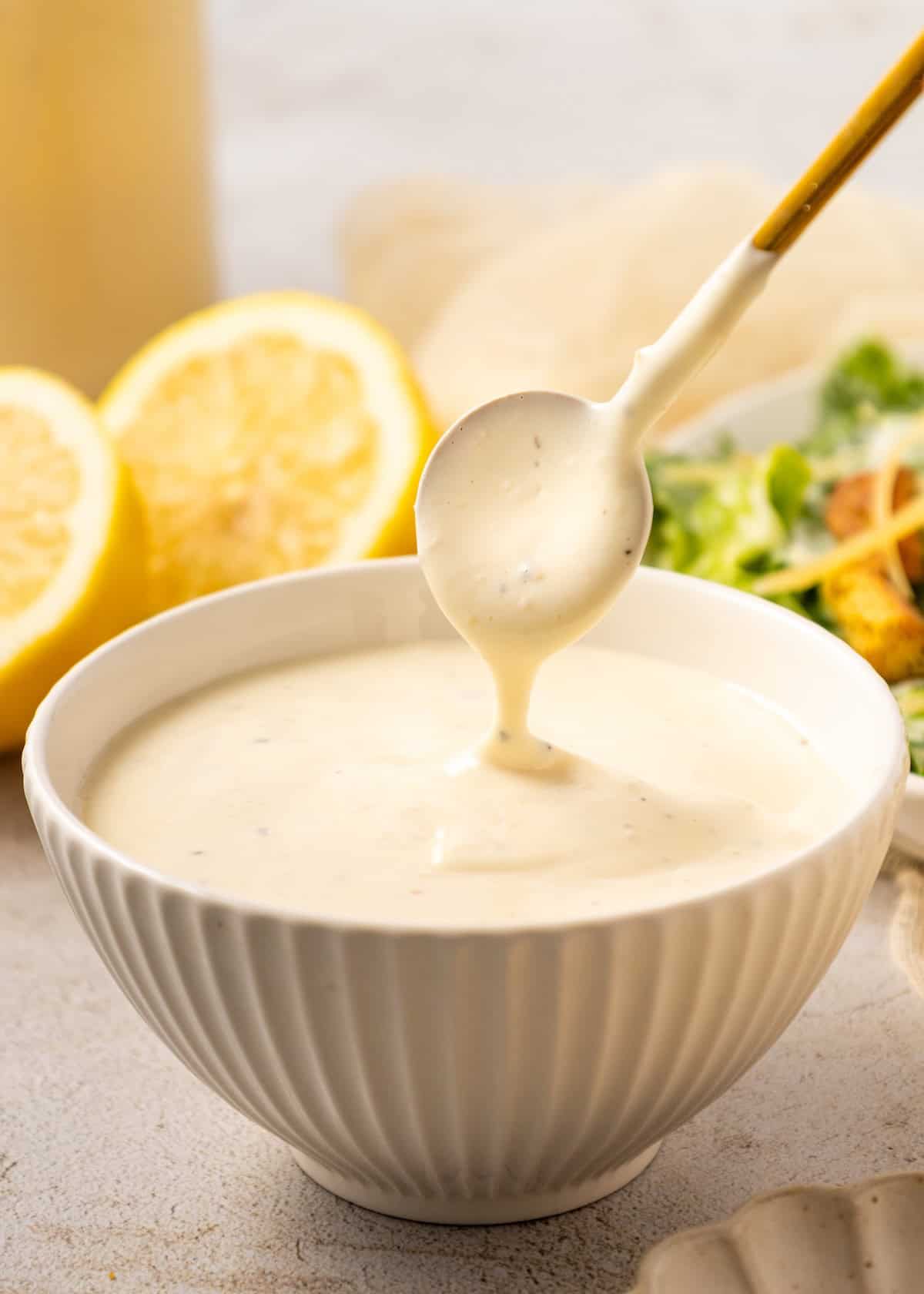 A spoon drizzling homemade Caesar dressing into a white bowl, with lemons and salad nearby.