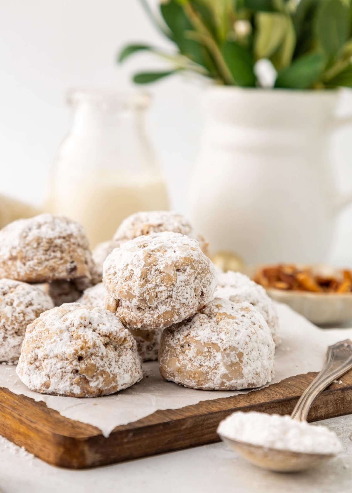 Pecan Snowball Cookies stacked on a wooden board with milk and greenery in the background.