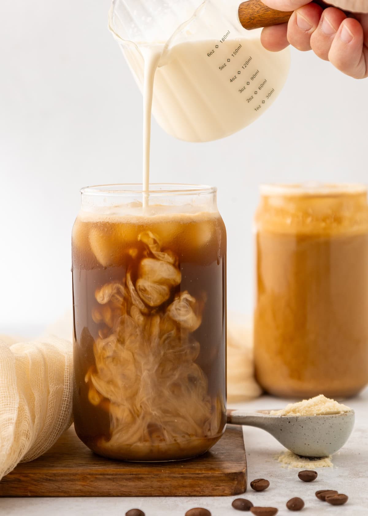 Cream being poured into a glass of Protein Iced Coffee, with beans and a jar in the background.