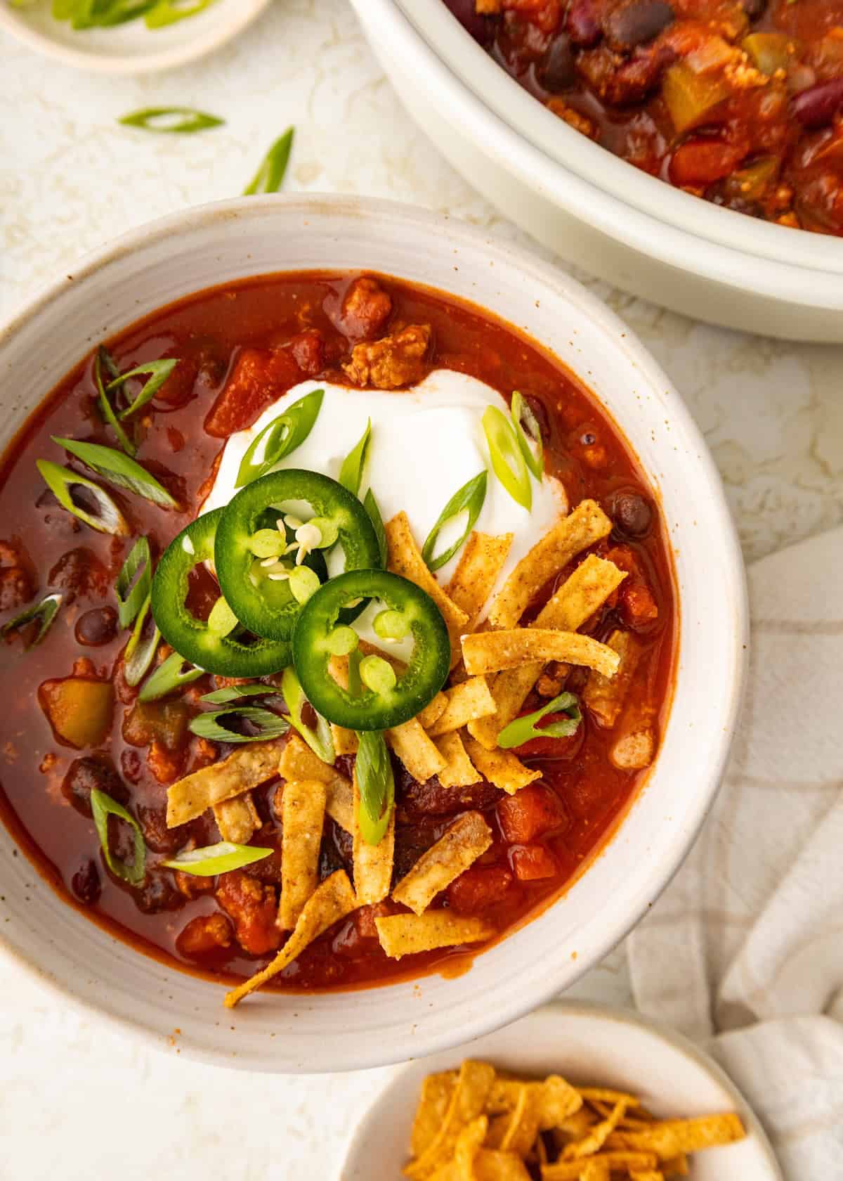 A bowl of ground turkey chili topped with sour cream, tortilla strips, and jalapeño slices.