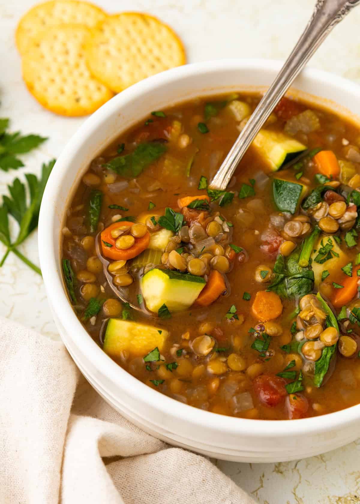 Bowl of Lentil Vegetable Soup with carrots, zucchini, parsley, and crackers on the side.