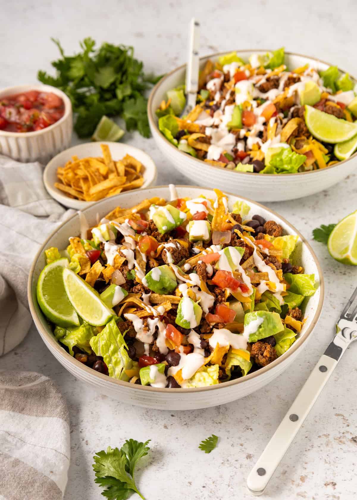 Two bowls of taco salad with lettuce, beans, fresh toppings, lime wedges, and creamy dressing.