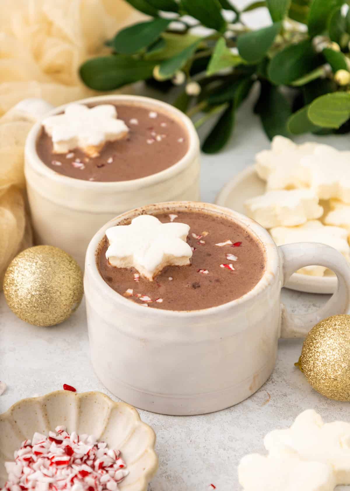 Two mugs of hot chocolate with snowflake marshmallows, surrounded by gold ornaments and greenery.