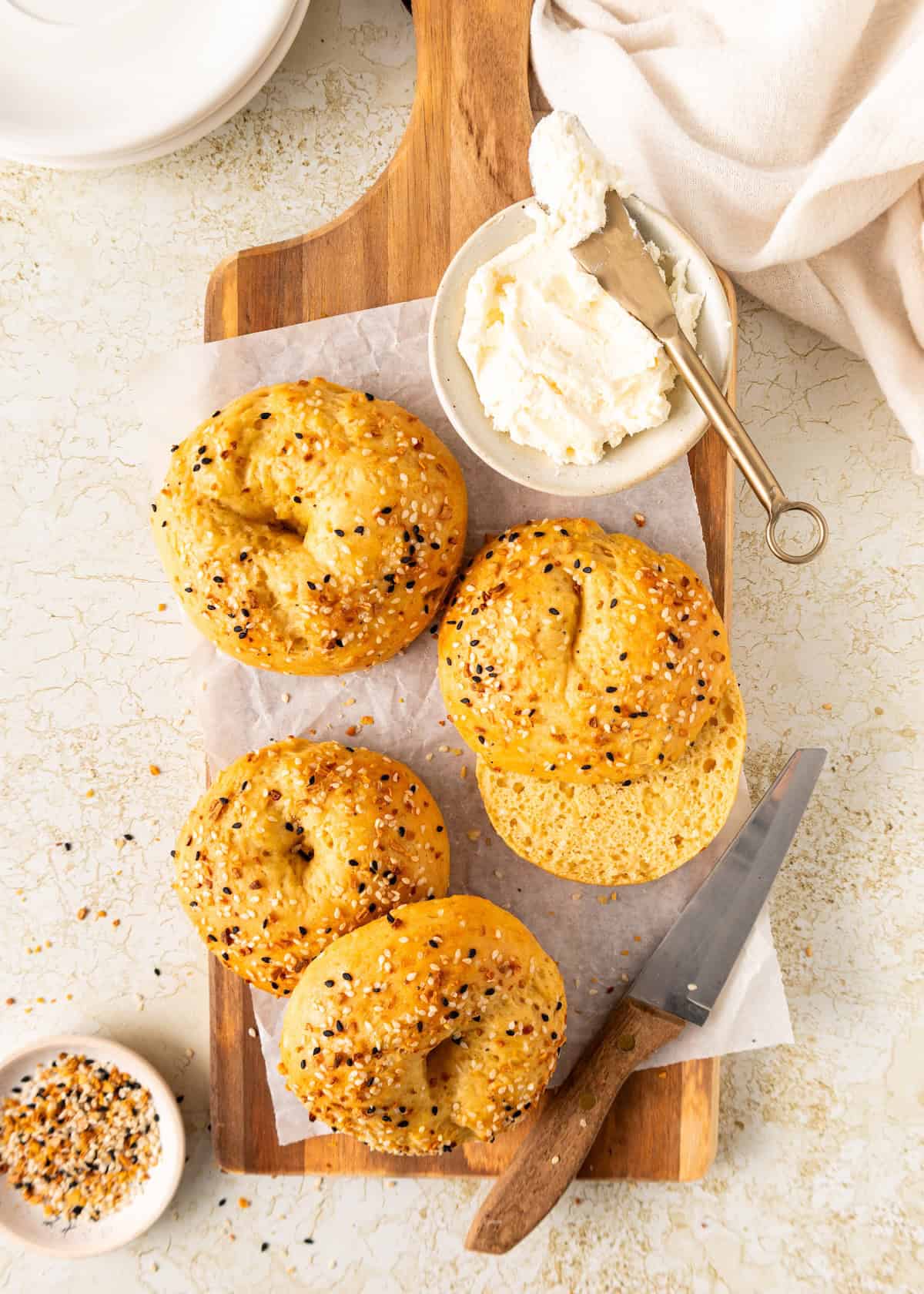 Four Greek Yogurt Bagels on a wooden board with cream cheese and a knife beside them.