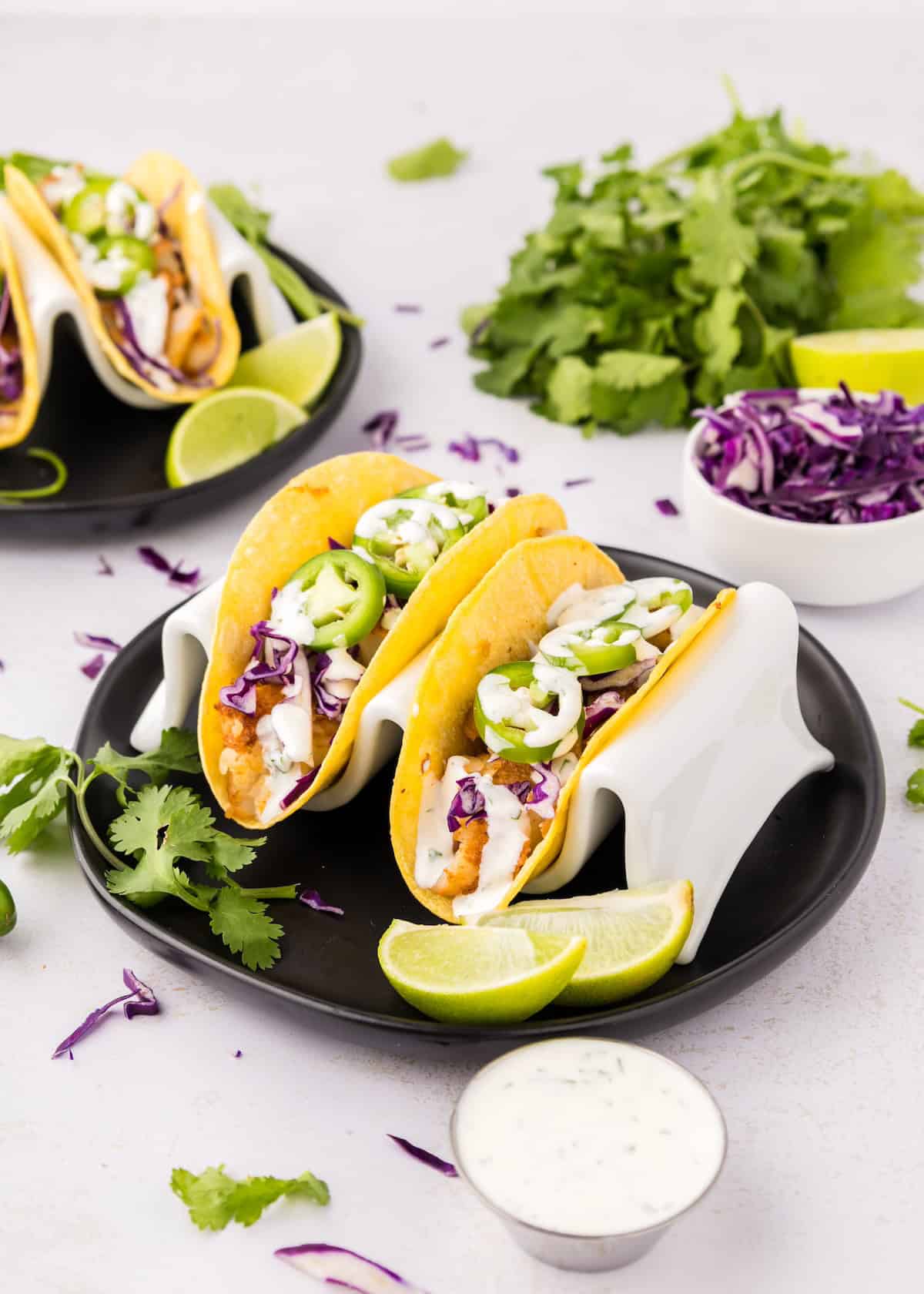 Two fish tacos with cabbage, jalapeños, and sauce, served with lime wedges on a black plate.