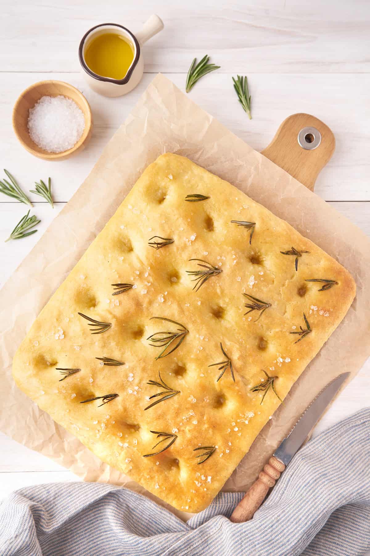 Square focaccia bread with rosemary on parchment, surrounded by salt, olive oil, and a knife.