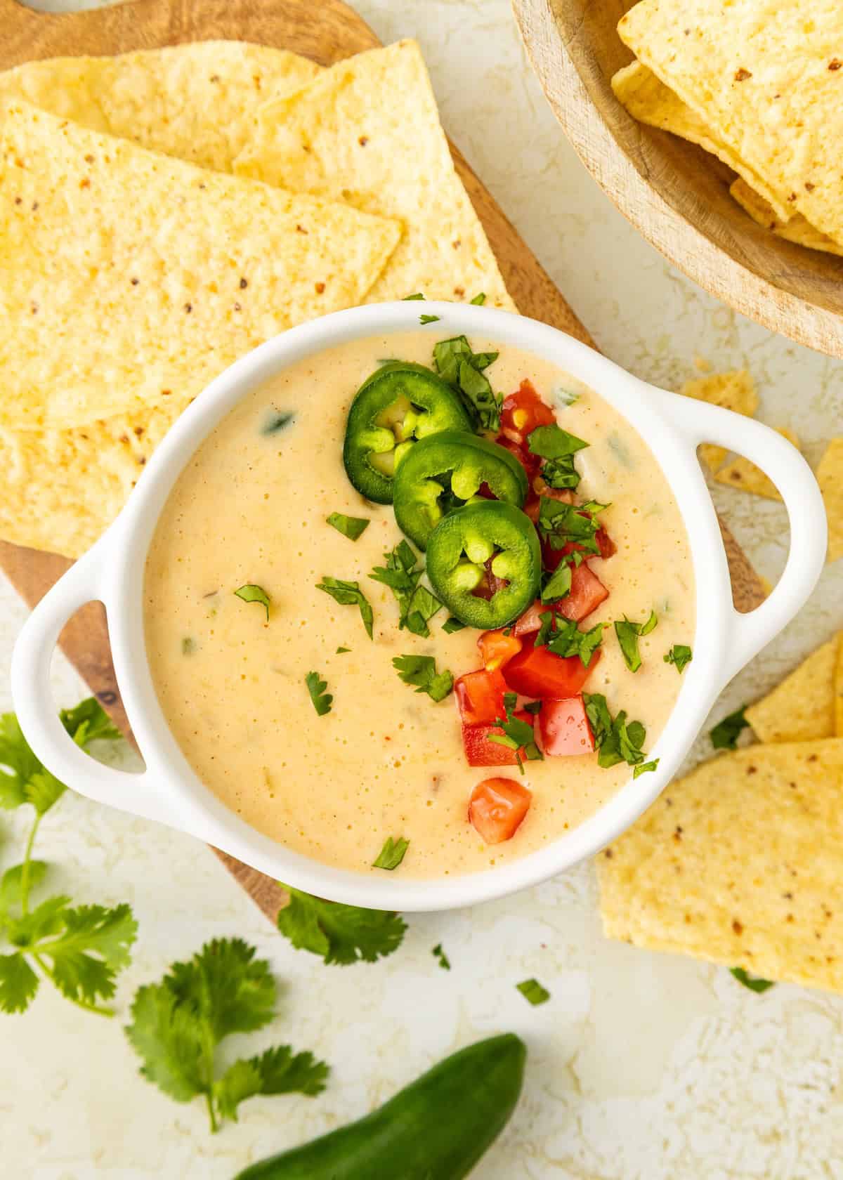 Bowl of queso dip topped with jalapeño, tomato, and cilantro, surrounded by tortilla chips.