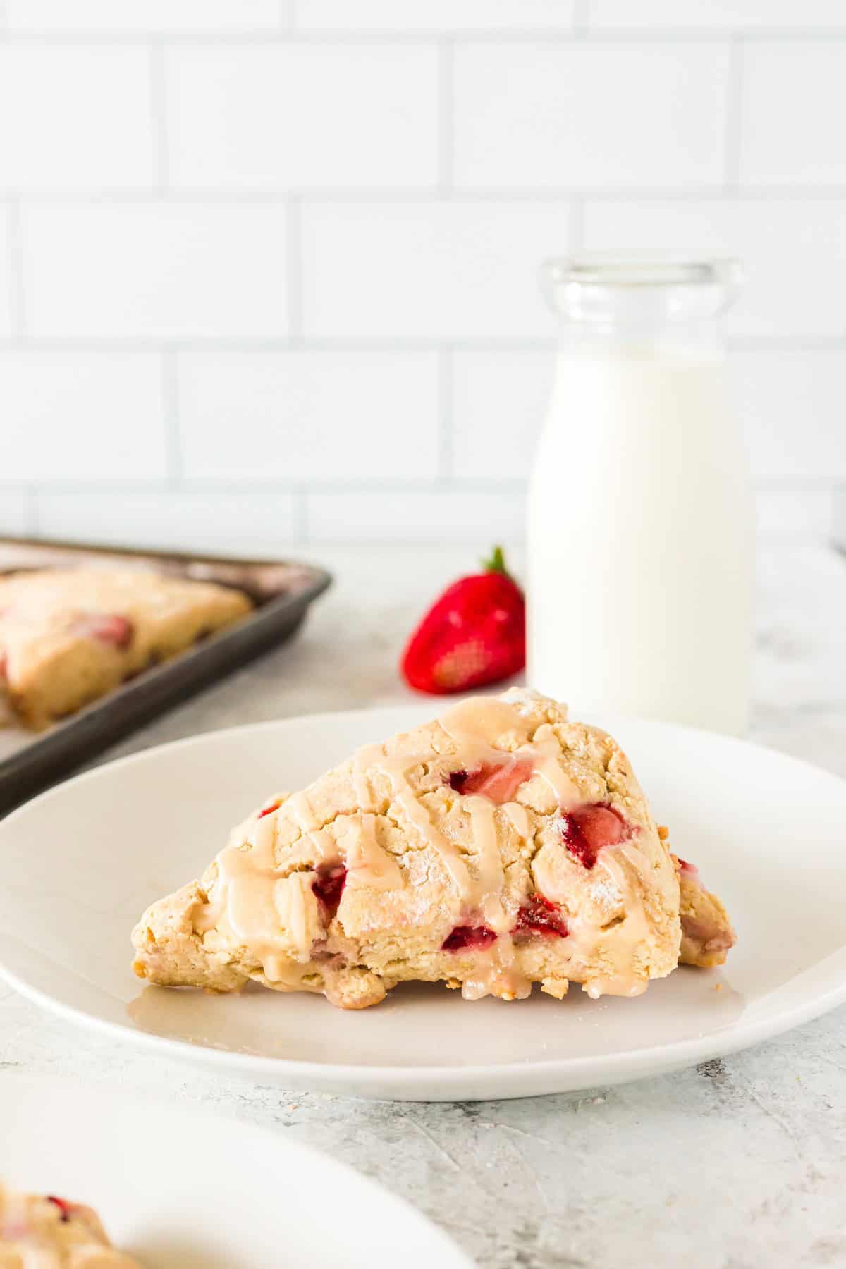 A strawberry scone on a plate with a bottle of milk and a strawberry in the background.