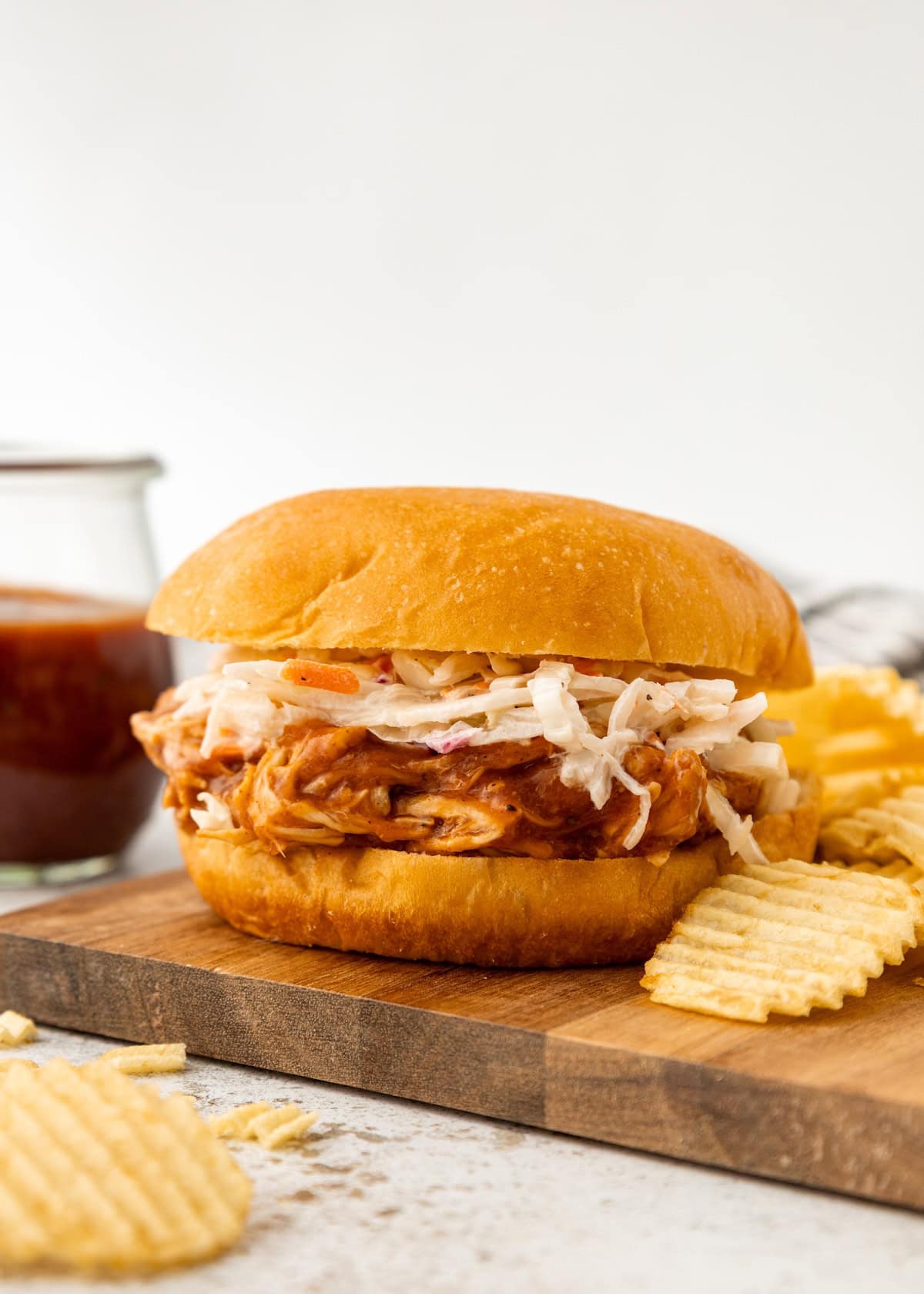 Pulled chicken sandwich with coleslaw on a bun, served with potato chips on a wooden board.